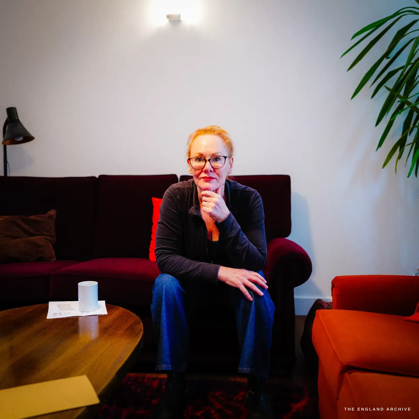 Grace Horne sits on a deep red sofa at home, hand to her chin, a potted palm beside her and a mug on the table.