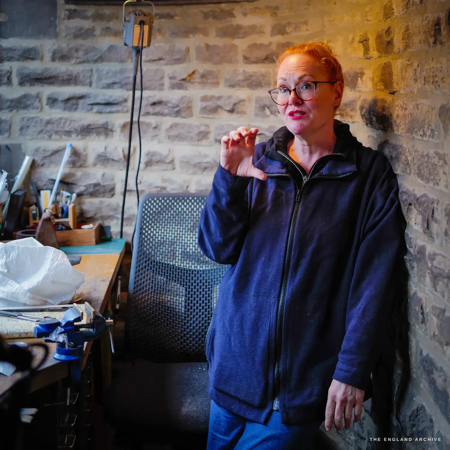 Grace Horne leans against the curved stone wall of the workshop, gesturing as she talks, the tool bench and vice to one side.