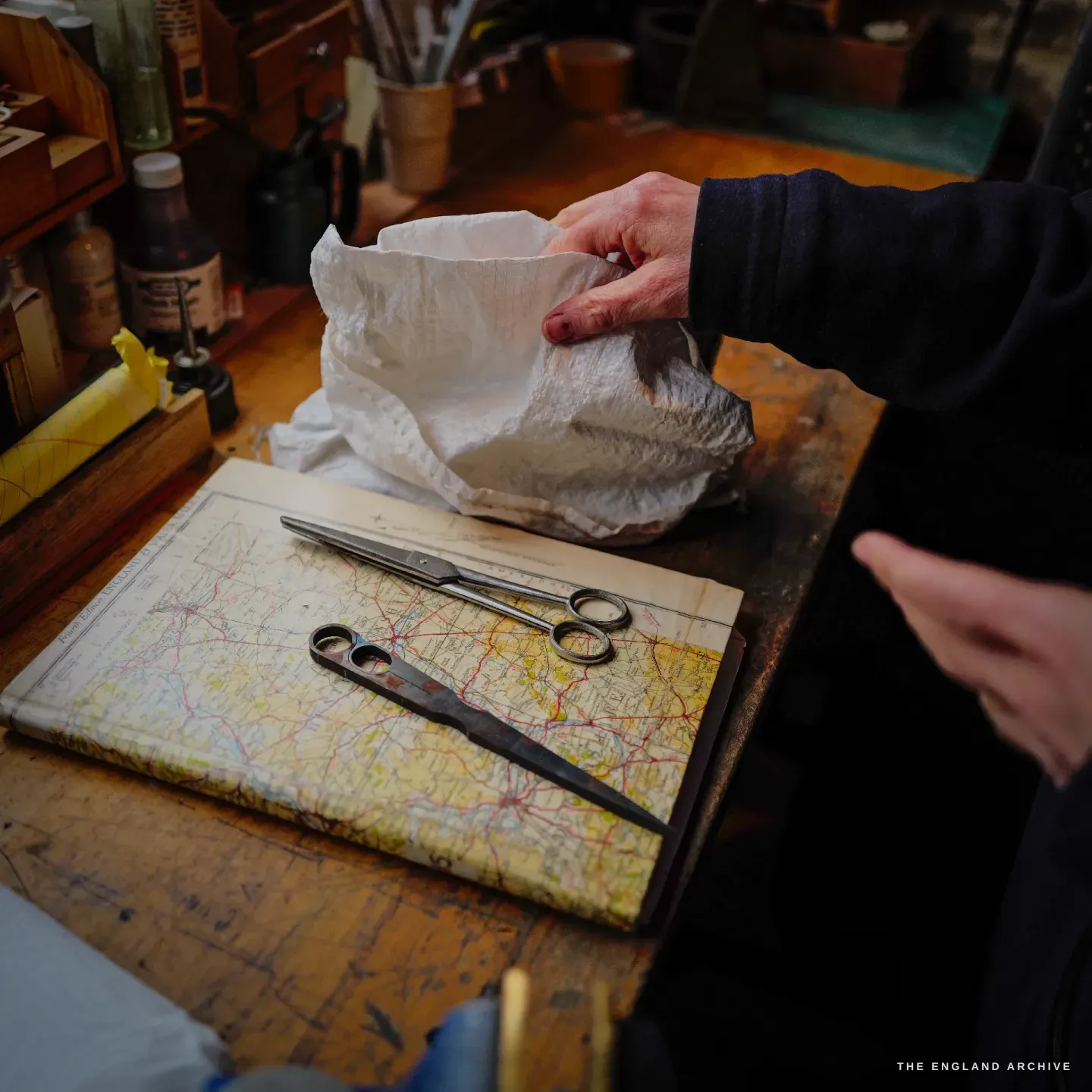 Two pairs of scissors rest on an old folded road map used as a bench cover, Grace Horne&rsquo;s hand lifting a cloth-wrapped parcel beside them.