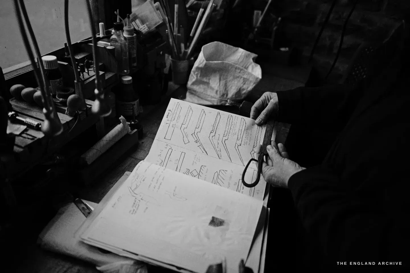 Grace Horne works at her bench over an open notebook filled with technical scissor drawings, a small pair of scissors in her hand.