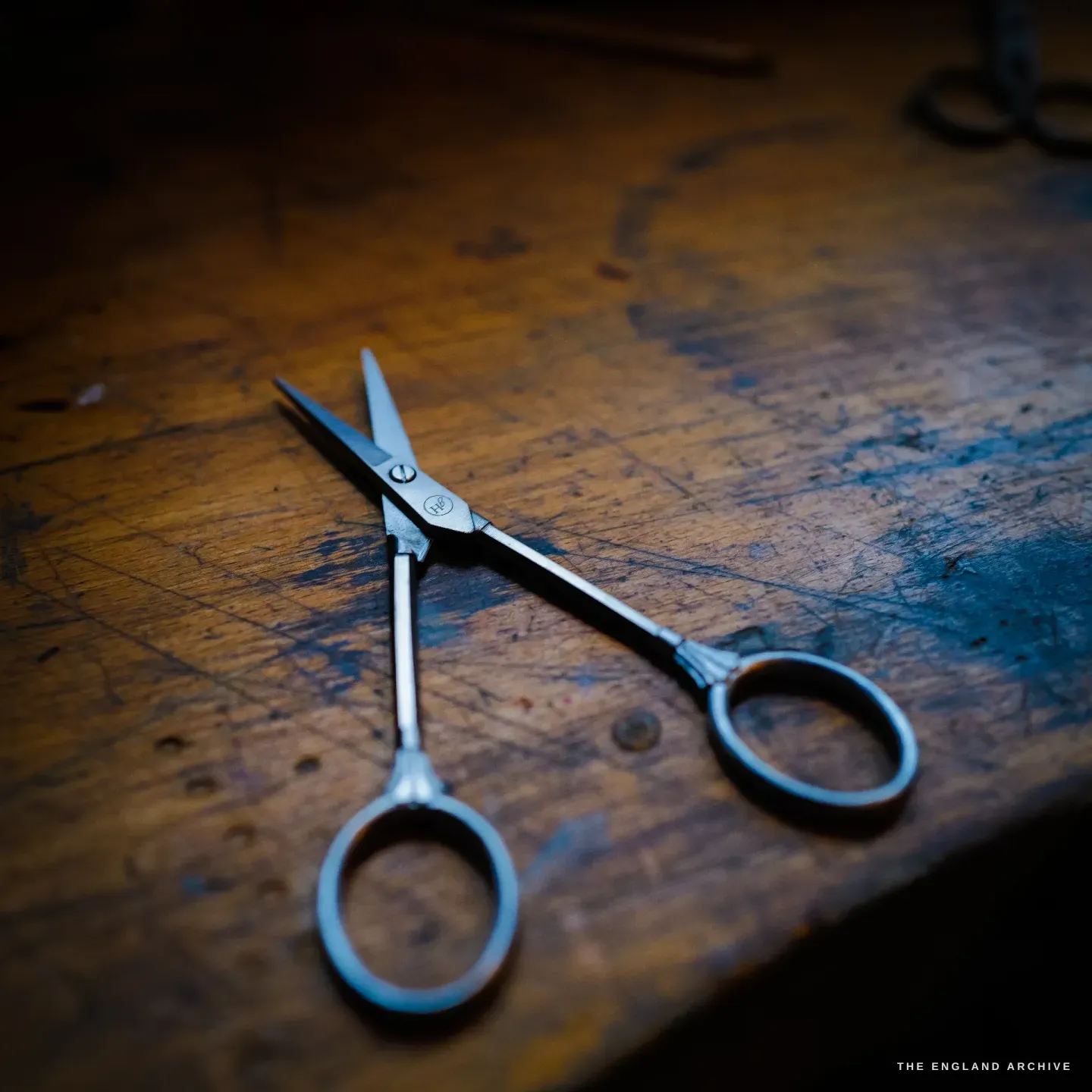A finished pair of polished scissors with a maker&rsquo;s stamp lies open on the worn wooden workbench in warm light, another forged pair behind.