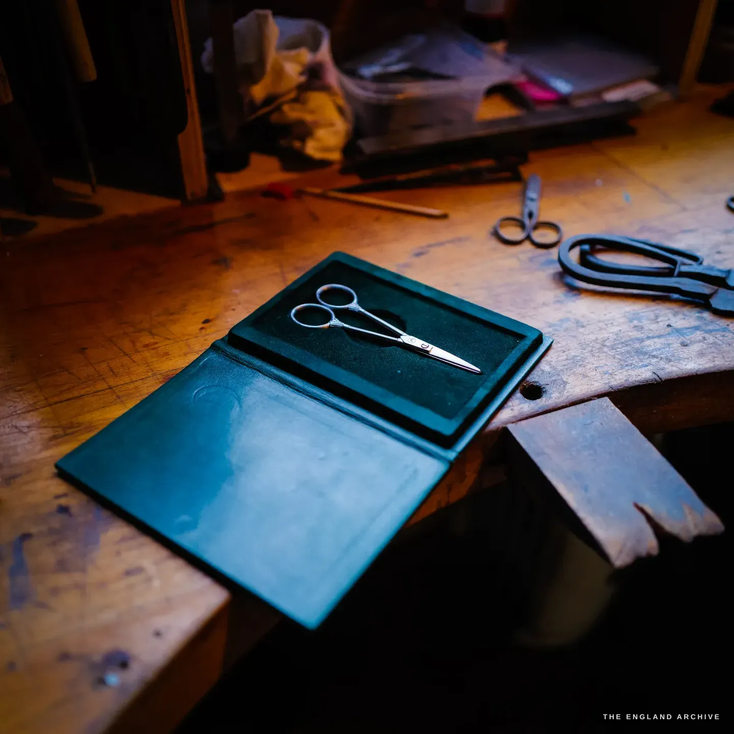 A finished pair of polished scissors displayed open on a dark green leather mat on the bench, other forged scissors beside it.