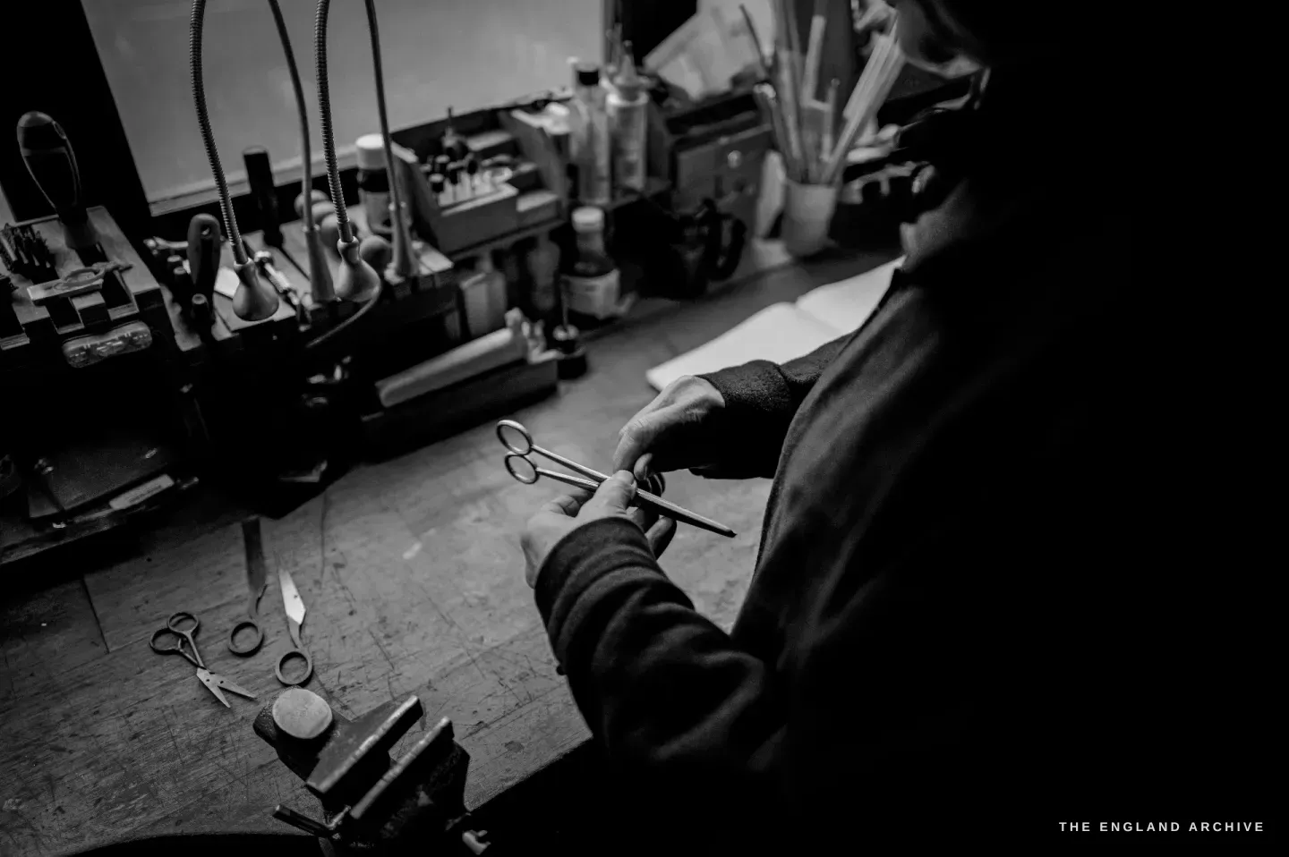 Over Grace Horne&rsquo;s shoulder, she works a small forged pair of scissors at the bench, more forged scissors and a vice in front of her.