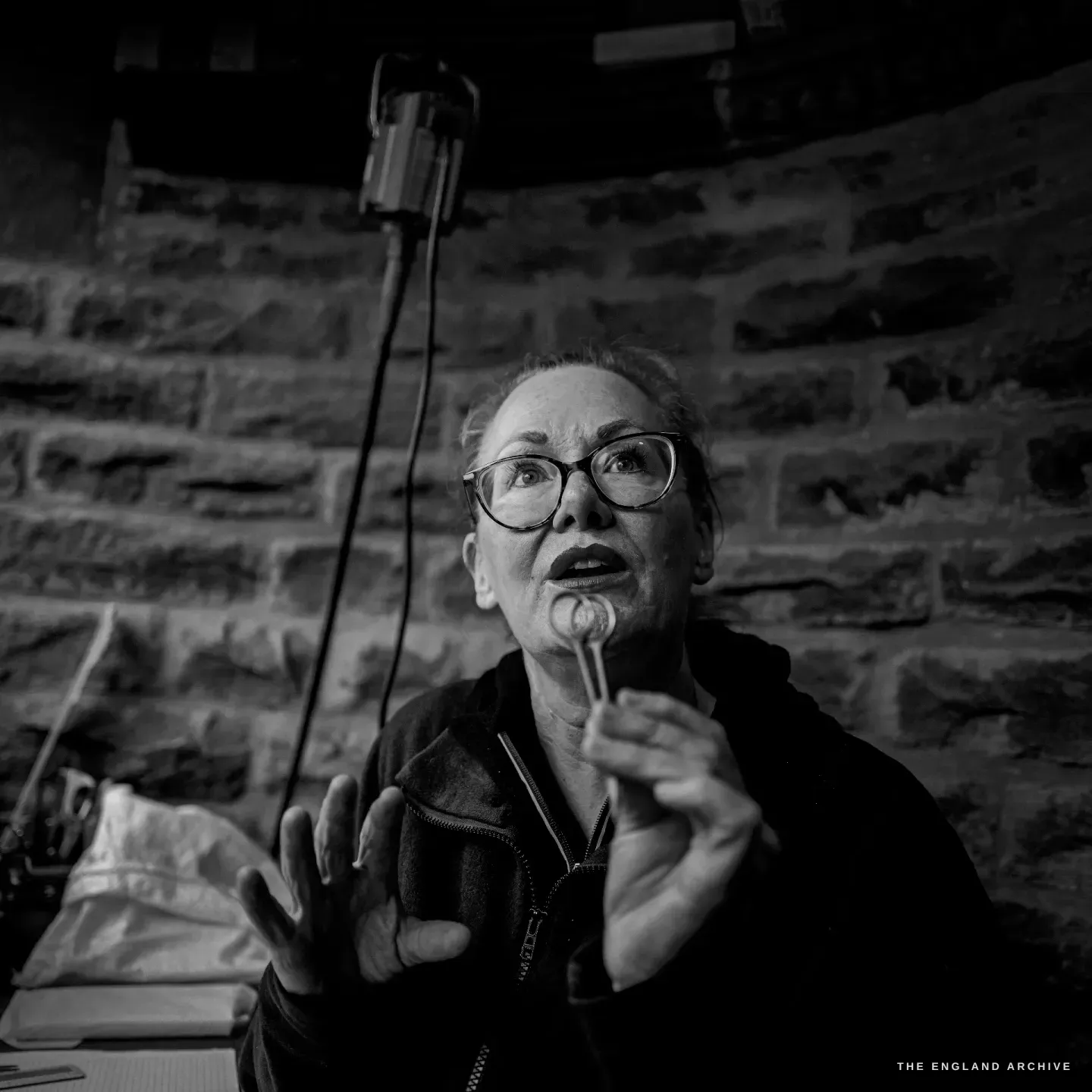 Grace Horne gestures with both hands as she explains something, a small pair of scissors held up, against the curved stone wall.