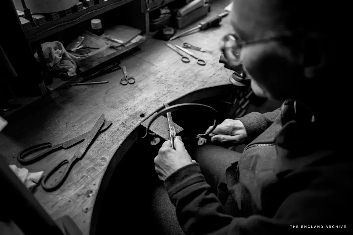 Over Grace Horne&rsquo;s shoulder she works the bow of a pair of scissors with a tool, several forged pairs laid out on the curved bench.