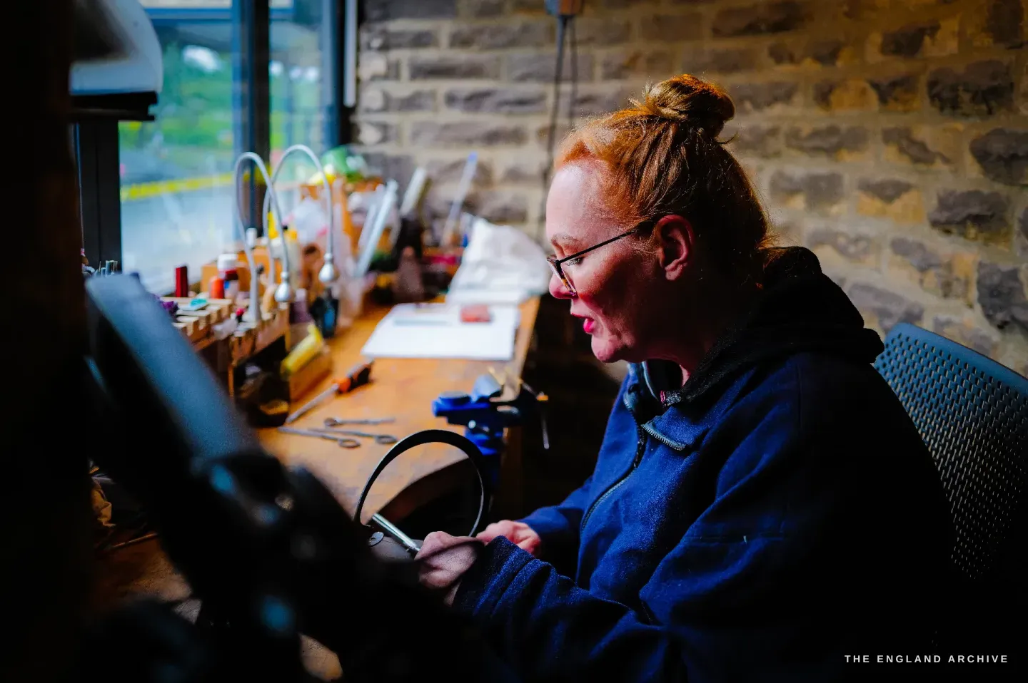Grace Horne sits at the bench by the window examining a scissor bow in her hands under the bench light, the stone wall behind.