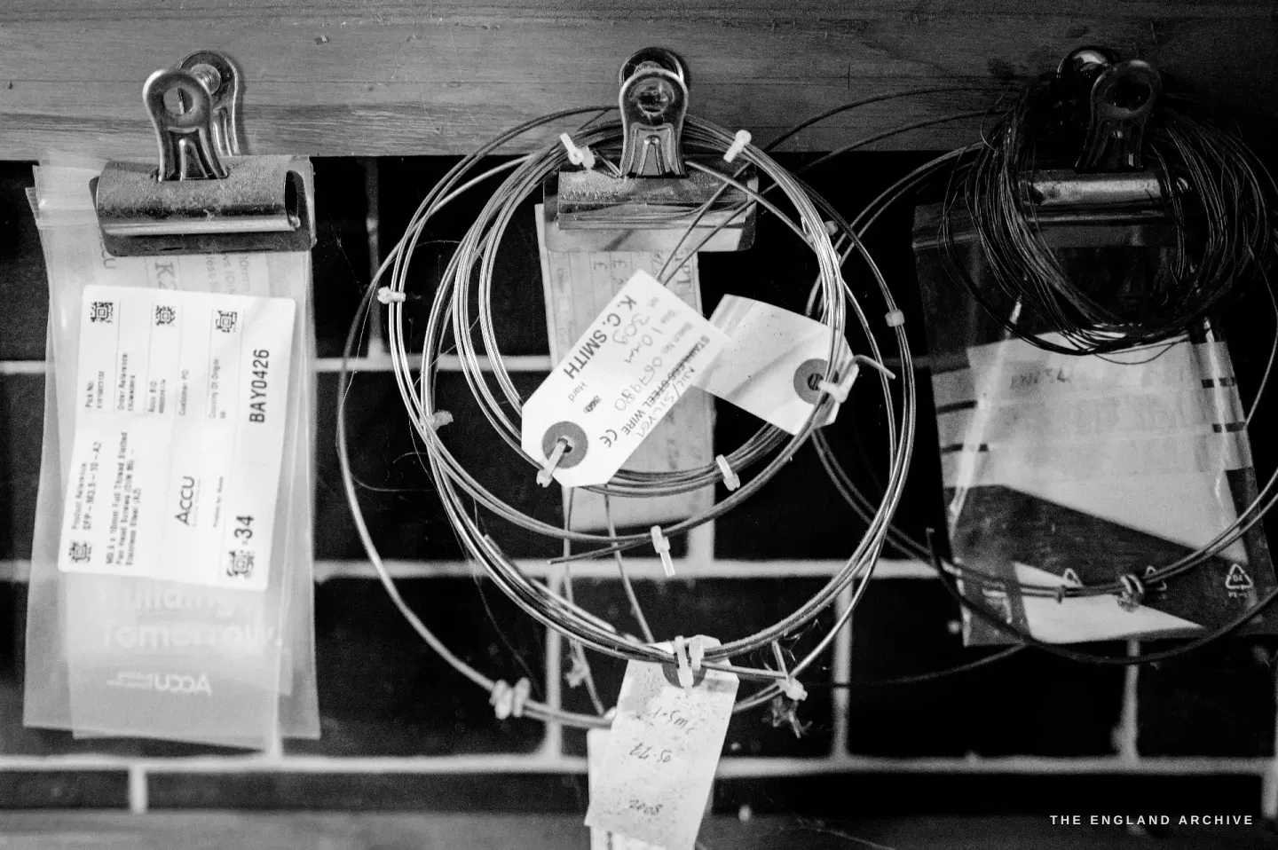 Coils of steel wire and paper tags clipped to a board with bulldog clips on the workshop wall - her stock and labelled notes.