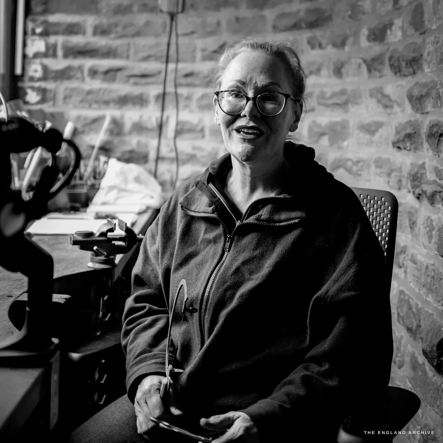 Grace Horne sits at her bench smiling warmly to the camera, a small pair of scissors in her hands, a microscope and vice beside her against the stone wall.