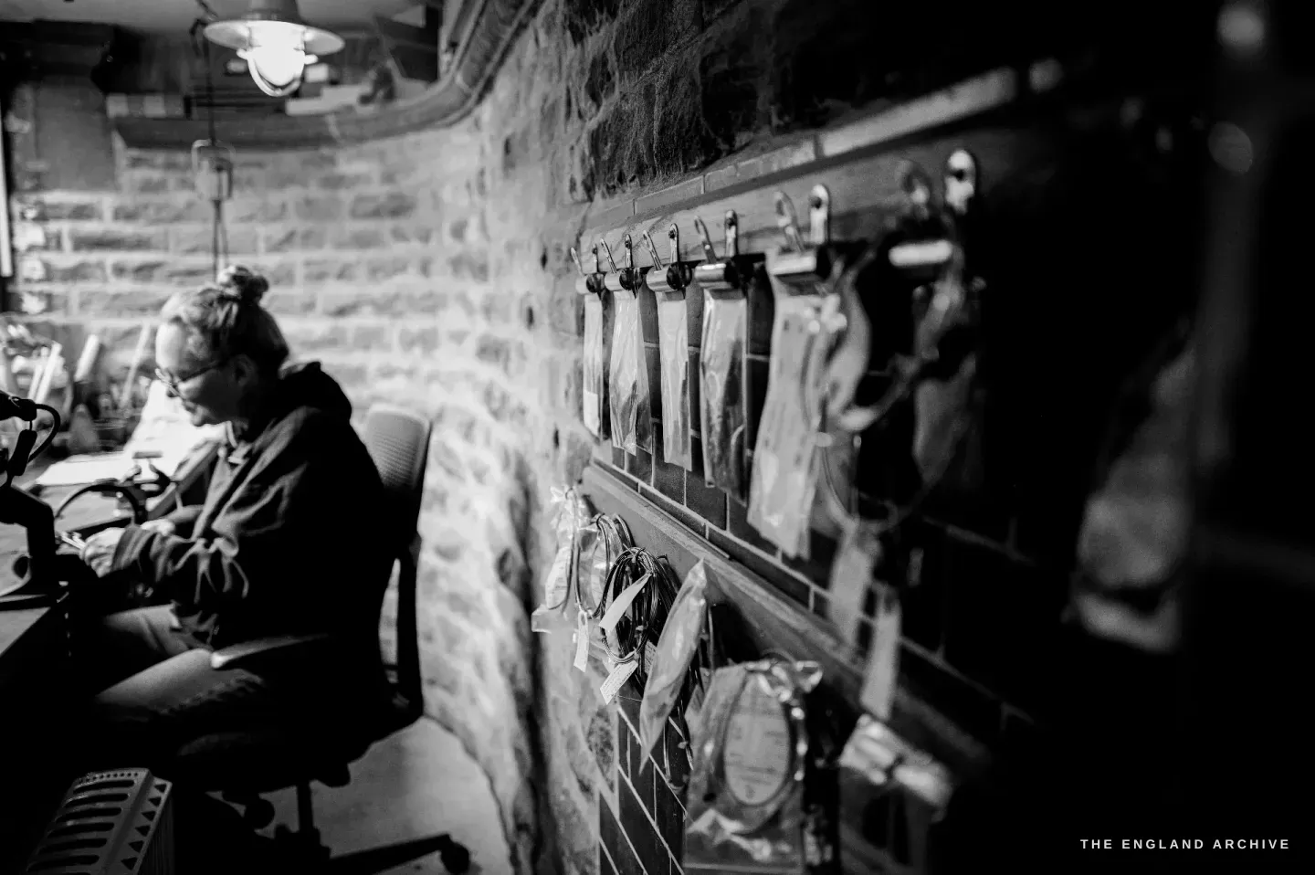 The workshop interior: Grace Horne works at the bench on the left while rows of scissors and tools hang from hooks along the brick wall on the right.