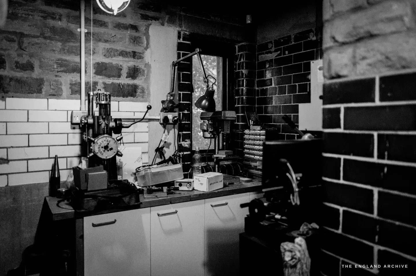 The workshop interior - a small drill press and machine on a bench beneath a window, the curved white-painted brick walls of the old building around it.
