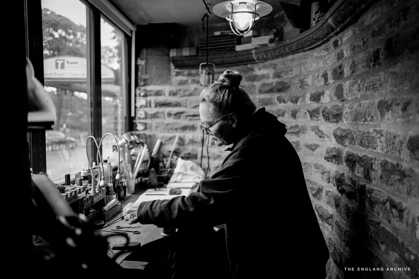 Grace Horne sits working at the bench beneath the window and an industrial lamp, the curved stone wall of the converted toilet curving around her.