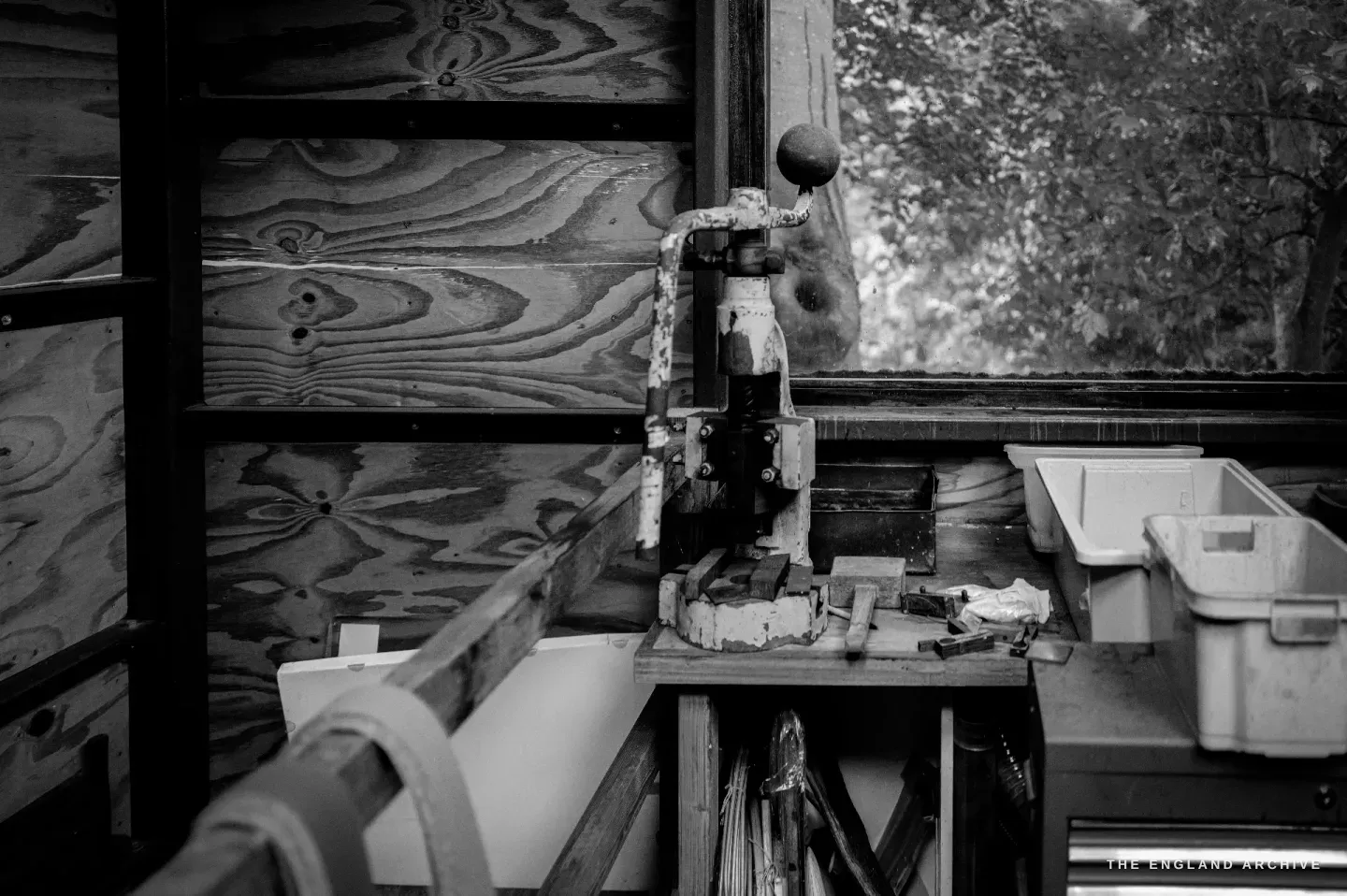 An old hand press mounted on a bench against a plywood wall in the workshop, trays beside it and greenery at the window.