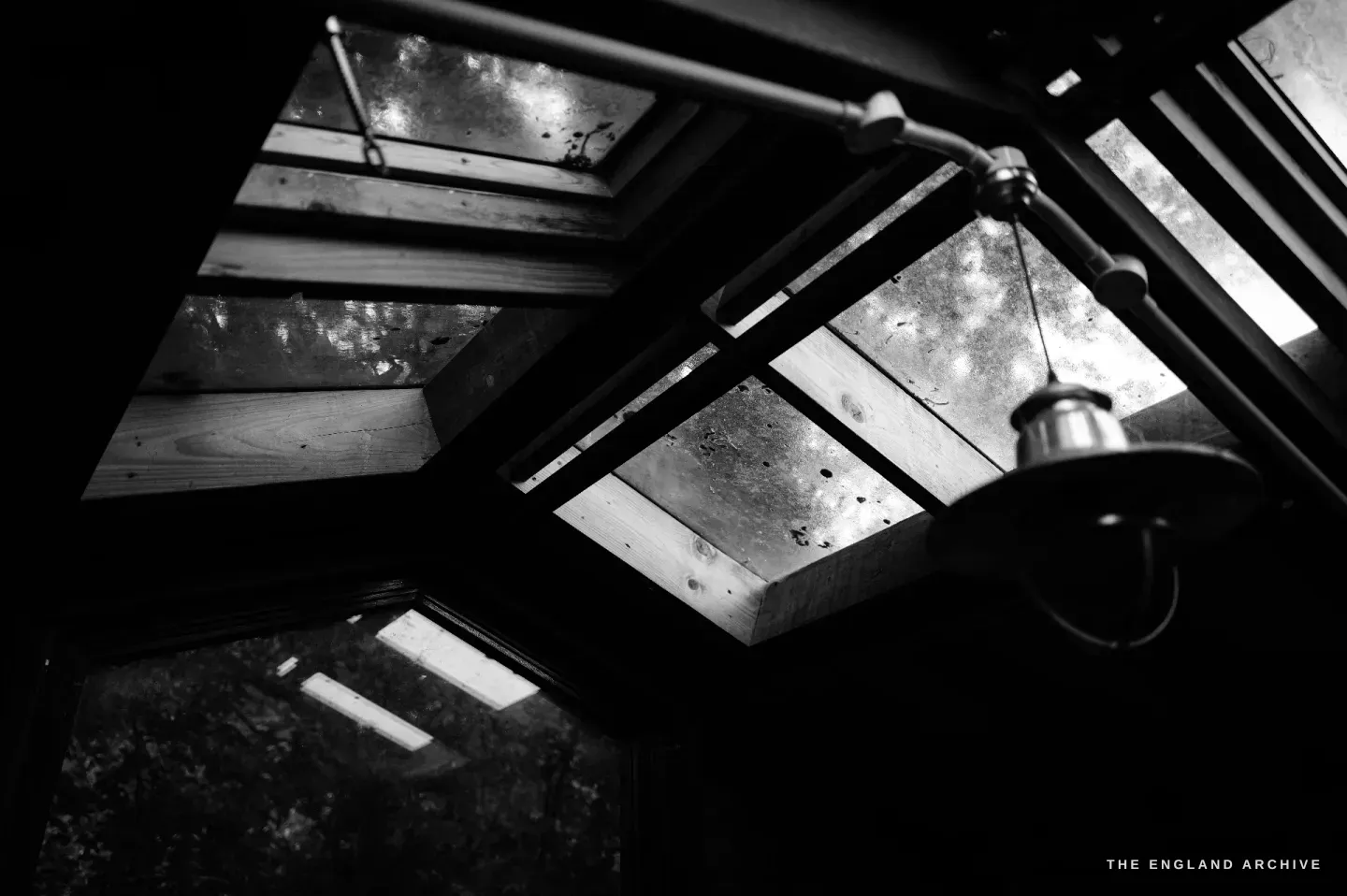 Looking up at the workshop&rsquo;s glazed roof - panes of old glass in a timber frame, an industrial pendant lamp hanging beneath.