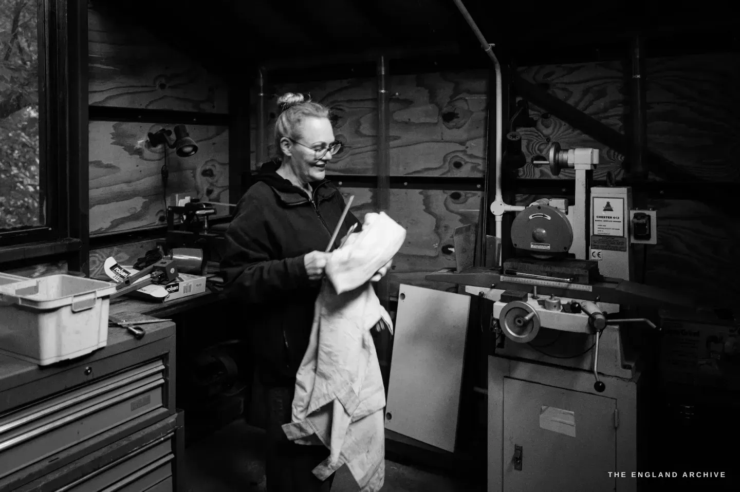 Grace Horne stands in a plywood-lined back room of the workshop holding a cloth, a bench grinder and tool chest beside her.