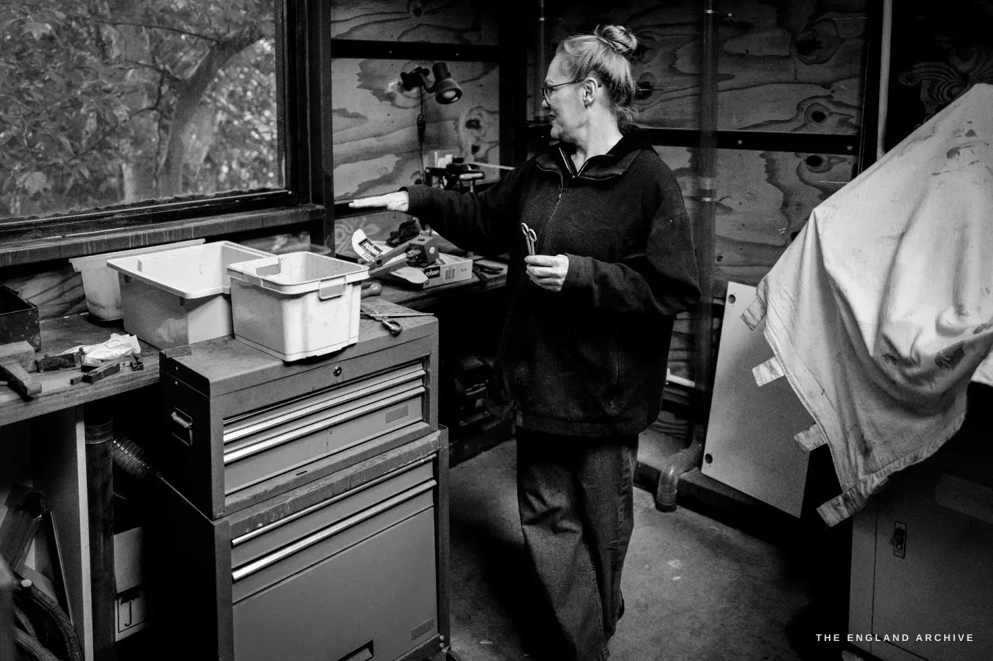 Grace Horne stands in the back machine room holding a scribe and small scissors, reaching toward a grey tool chest by the window.