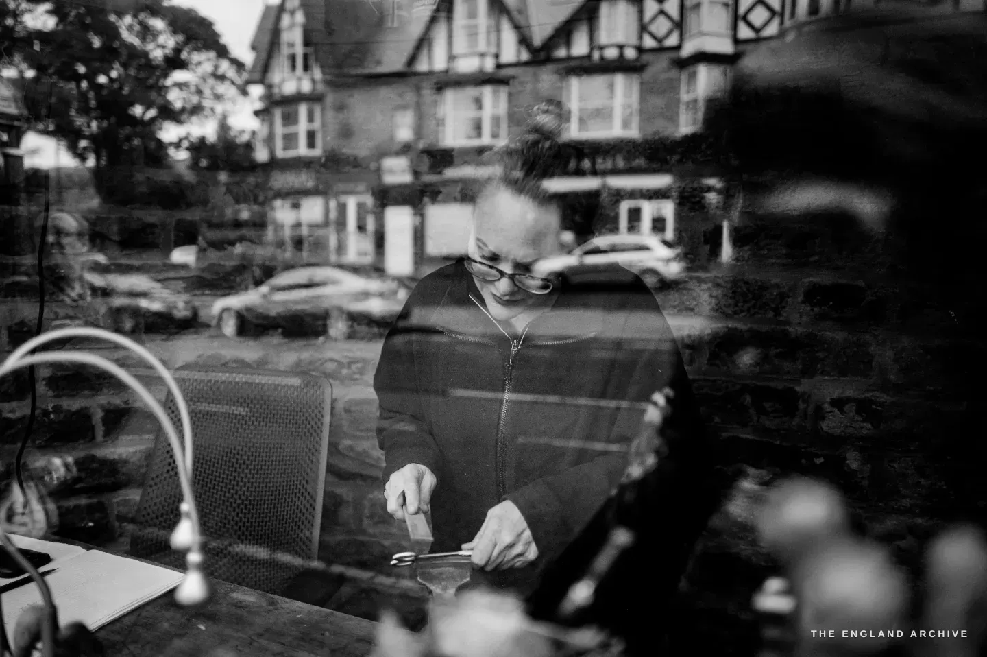 Grace Horne seen through the workshop window at her bench, the terraced street and parked cars reflected across the glass over her.