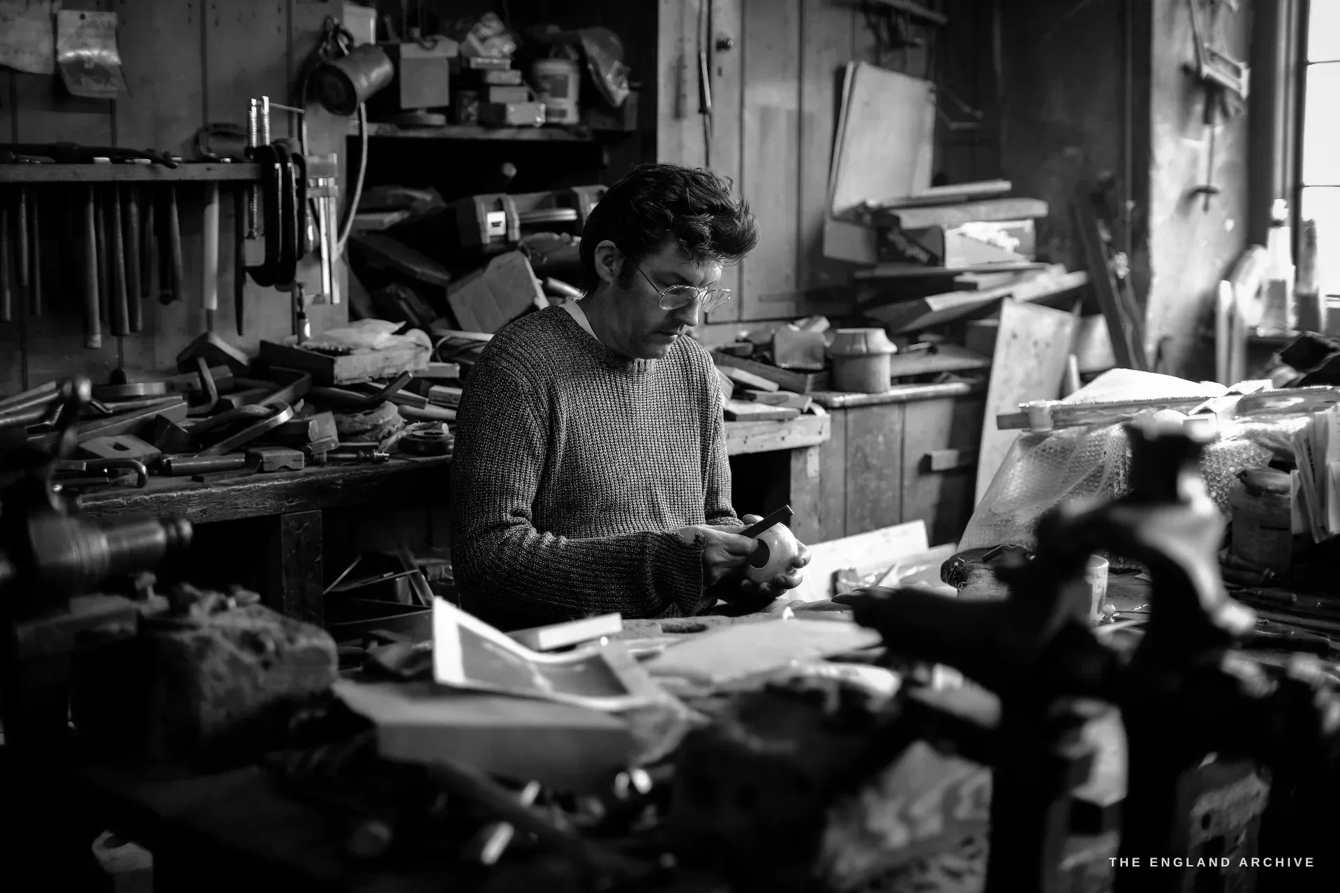 A dark-haired man in a wool jumper sits at a cluttered workbench, examining a small silver piece between his fingers, racks of tools and offcuts around him in the dim workshop.