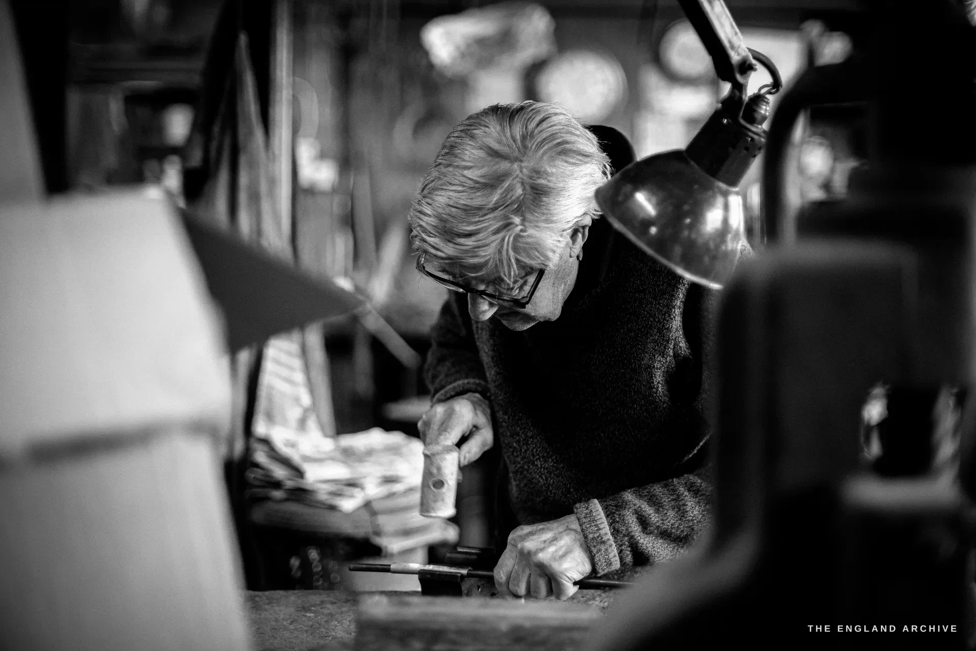 A white-haired silversmith bends low over his bench under an angled lamp, gripping a small punch against the work in close concentration.