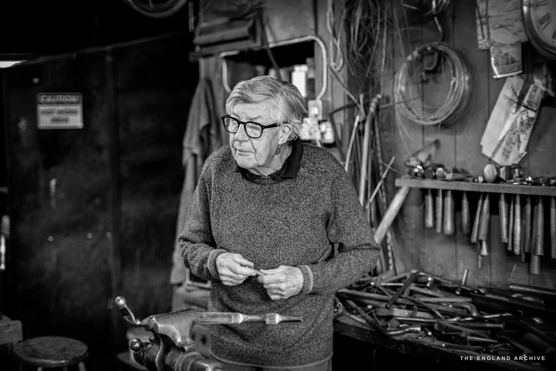 David Hart, bespectacled and white-haired in a heathered jumper, stands at his bench holding a small silver piece, a wall of hanging hand tools and metal hoops behind him.