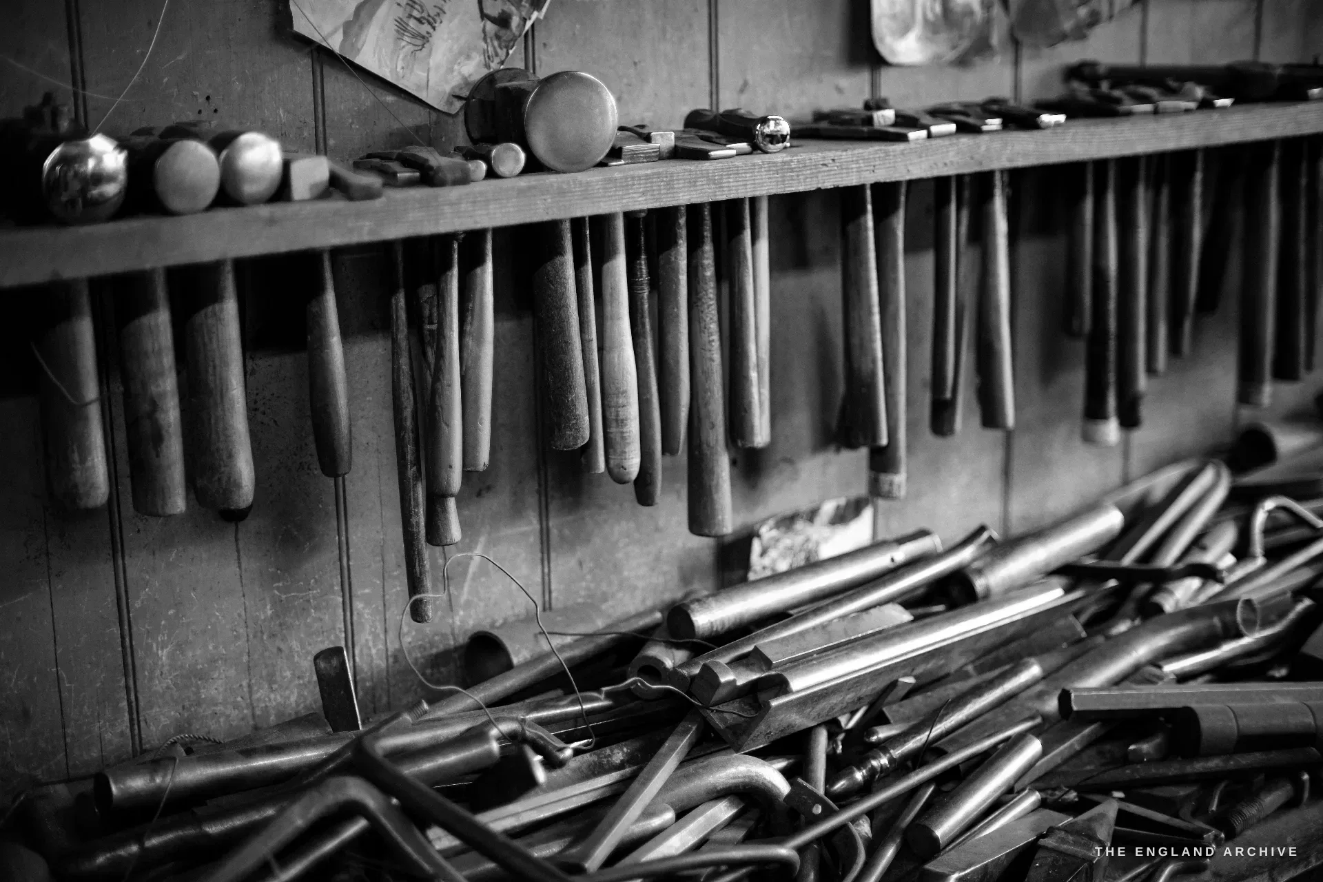 A long wall rack of hammers and hanging hand tools above a heaped tray of curved steel stakes and rods, the boarded wall of the workshop behind.