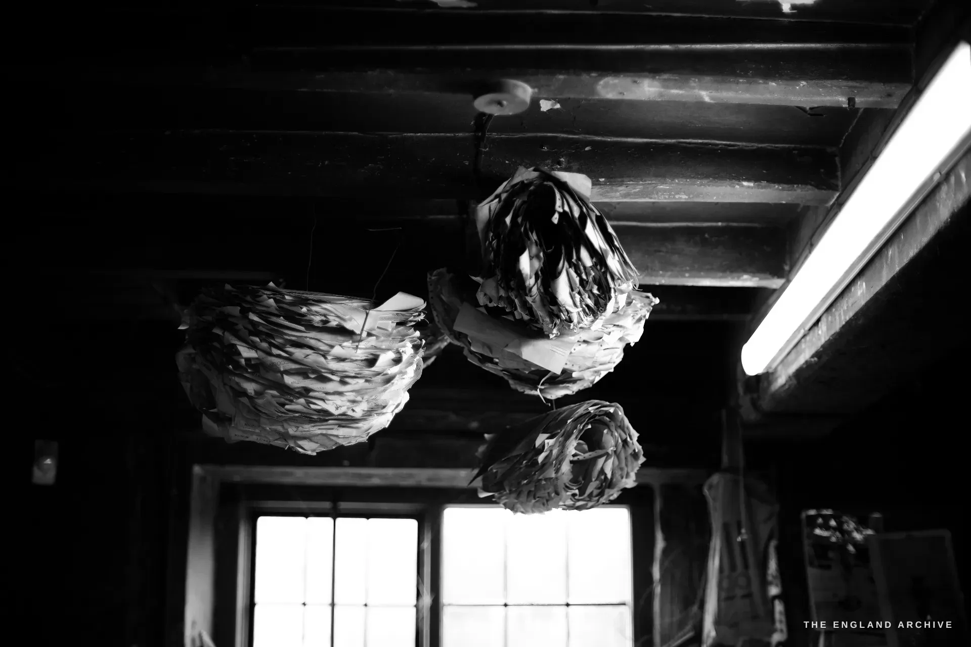 Looking up to the workshop ceiling beams where three large balls of spiked paper receipts hang, a strip light glaring at the right.