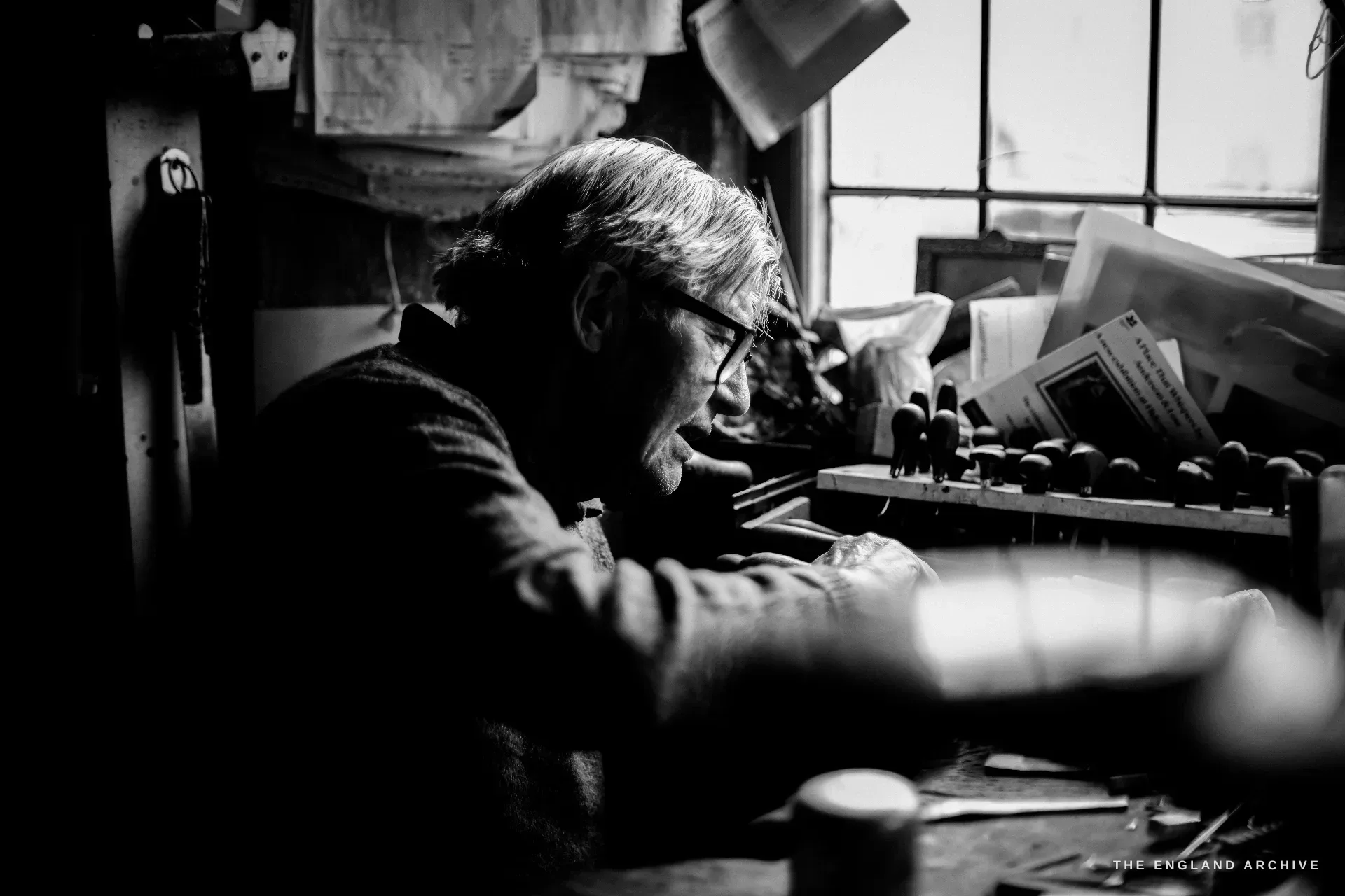 David Hart in profile at his window-lit bench, leaning on folded arms beside a rack of doming punches, papers pinned on the wall above.