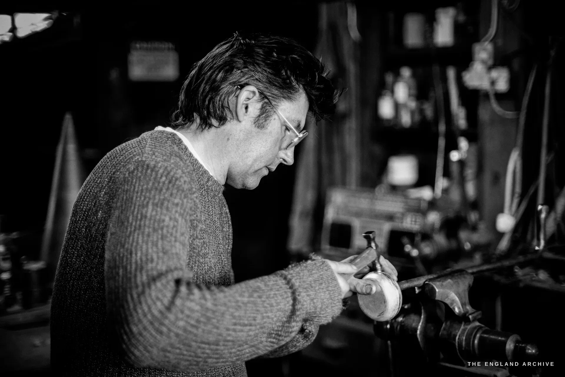A dark-haired bespectacled man in a wool jumper works a silver piece against a polishing spindle at the bench, side-lit by the window.