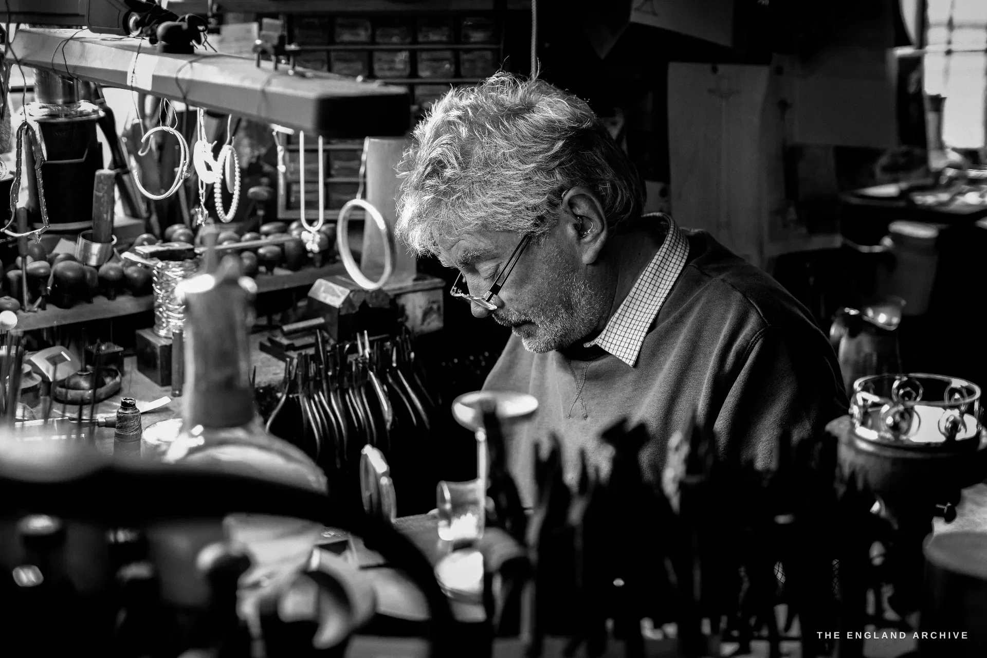 A grey-haired bespectacled silversmith bends over his bench amid a dense foreground of finished silverware - candlesticks, cups and trophies catching the light.