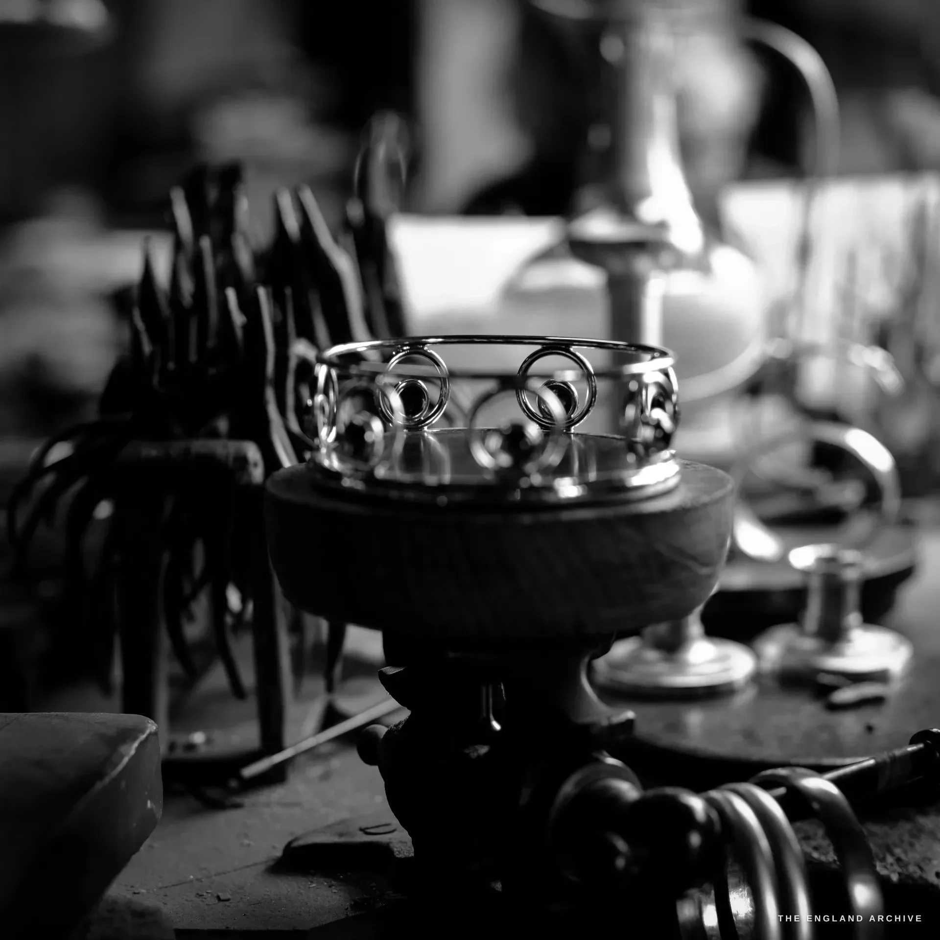 A polished silver pierced-gallery dish set on a wooden doming block, glittering against a soft-focus clutter of tools and silver.