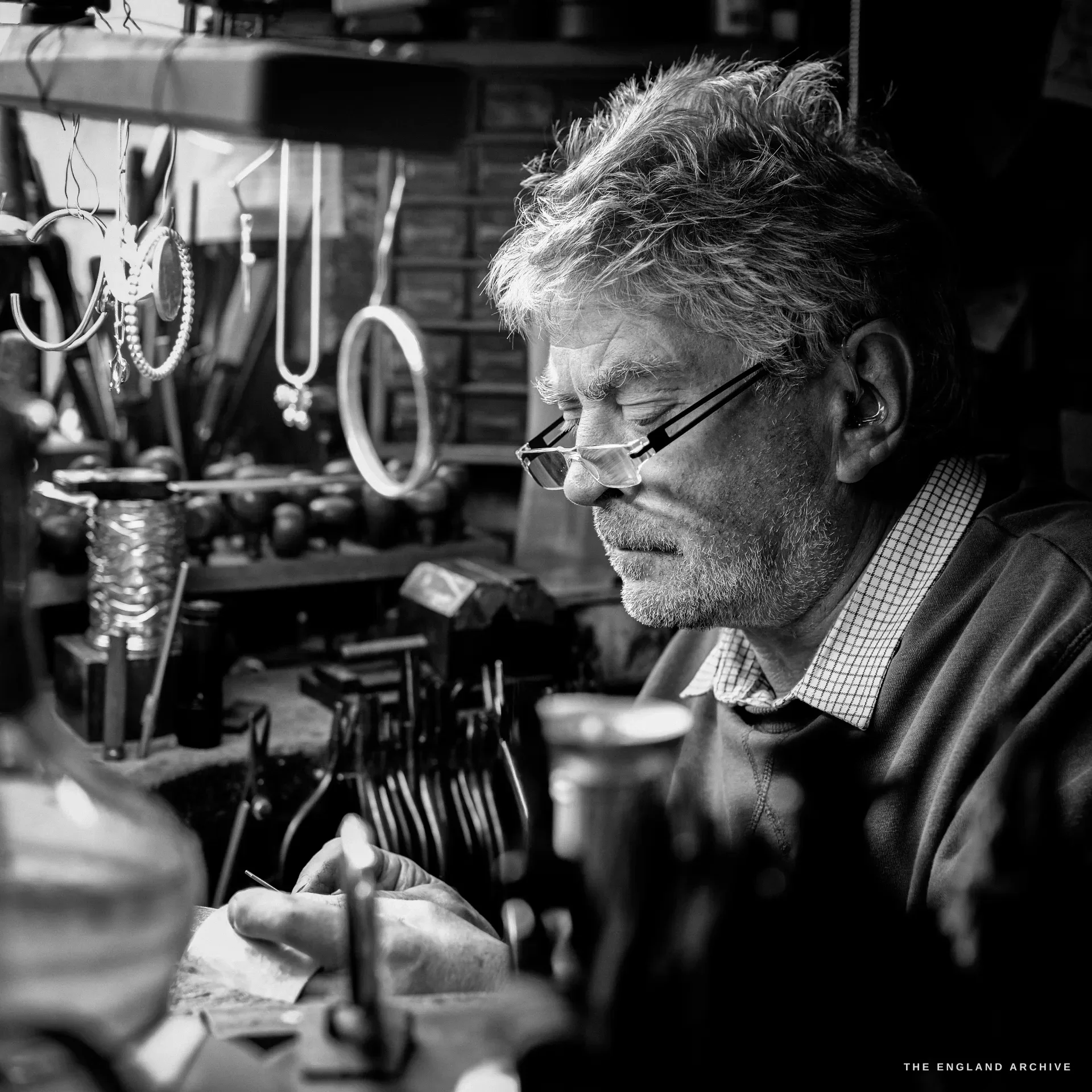 A close three-quarter view of a grey-haired bespectacled silversmith bent in concentration over his bench, racks of bangles and tools softly behind.
