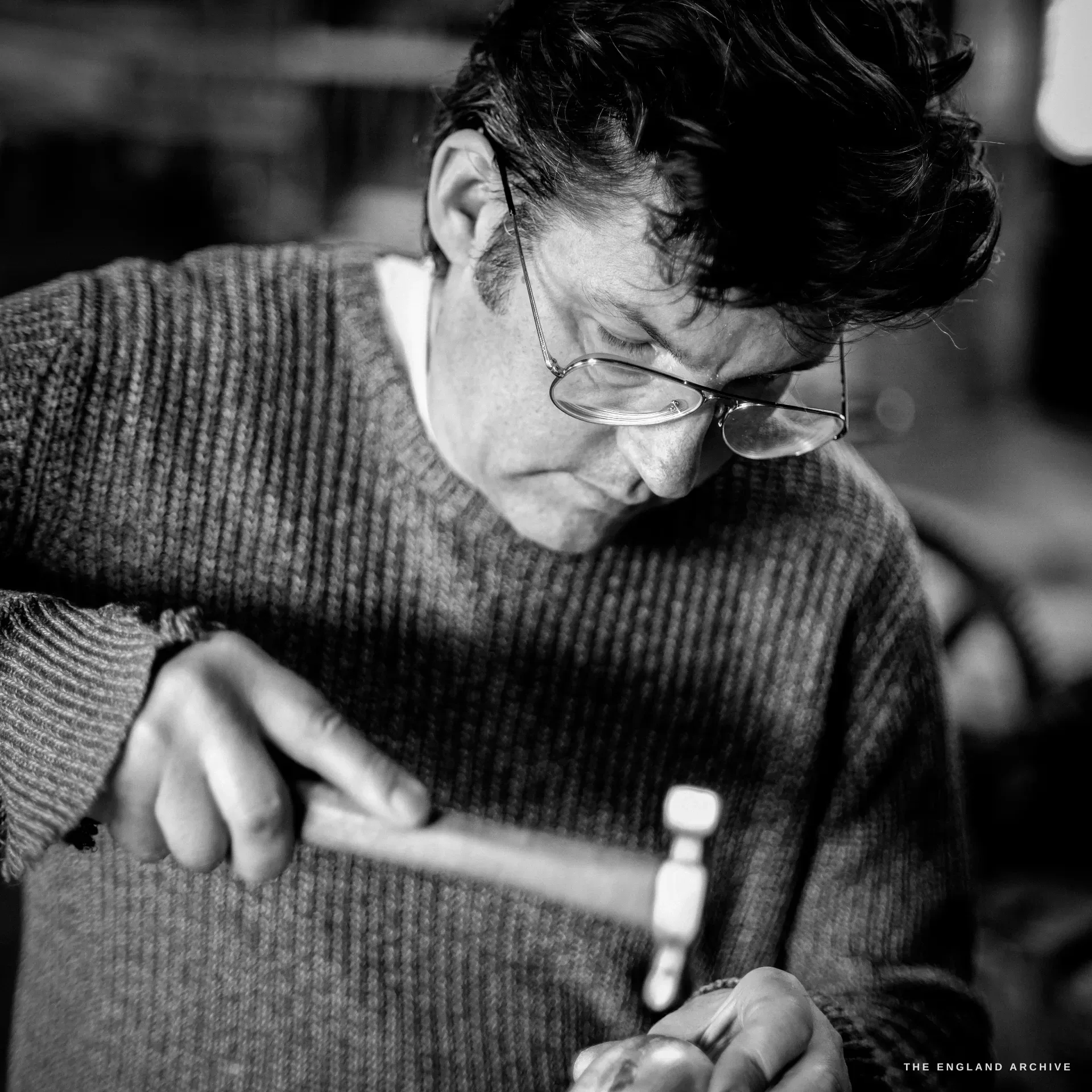 A dark-haired bespectacled man in a ribbed jumper bent over his work, holding a hammer and a small silver piece, eyes down in focus.