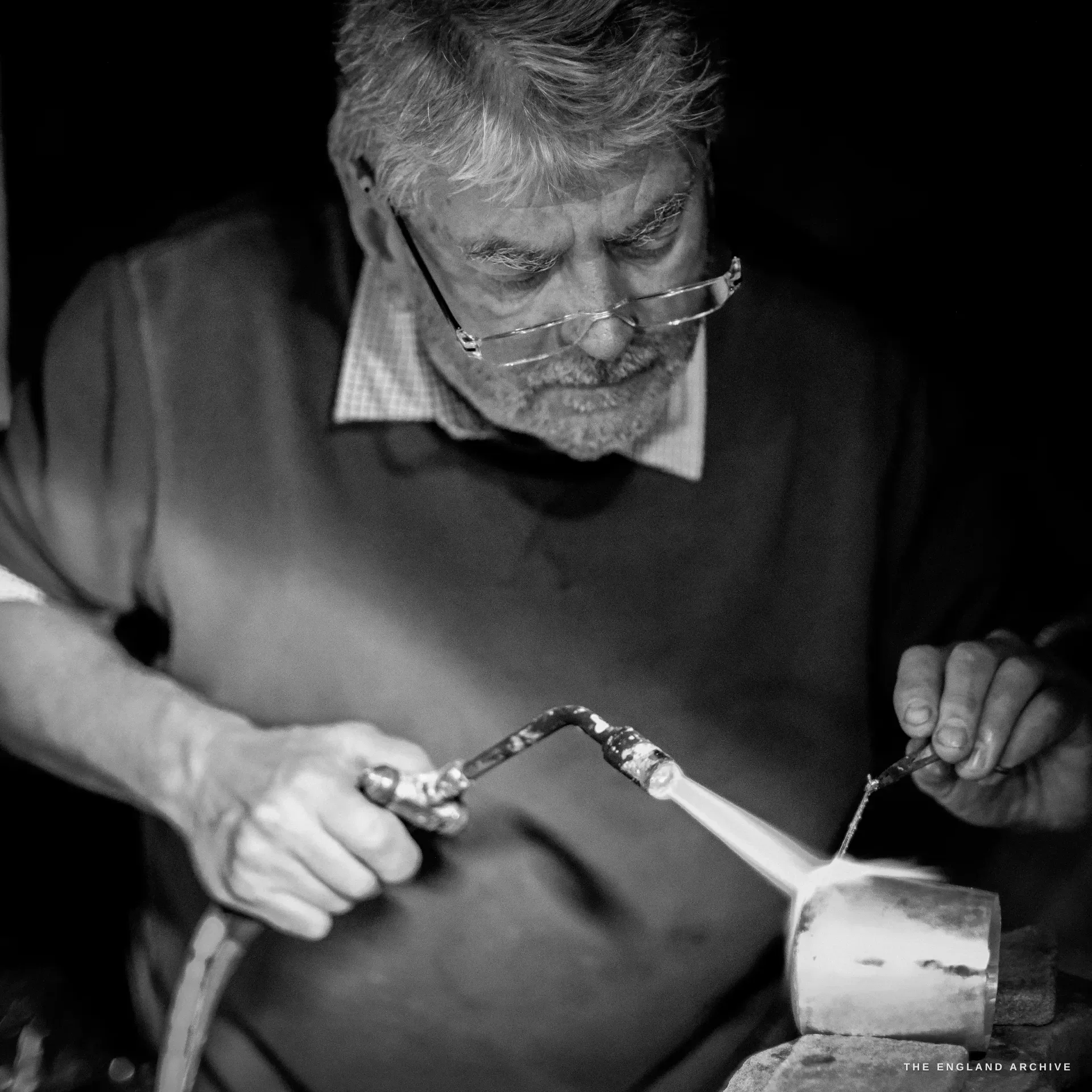 An elderly bespectacled silversmith plays a gas blowtorch over a silver workpiece held in tongs, the flame lighting his face.