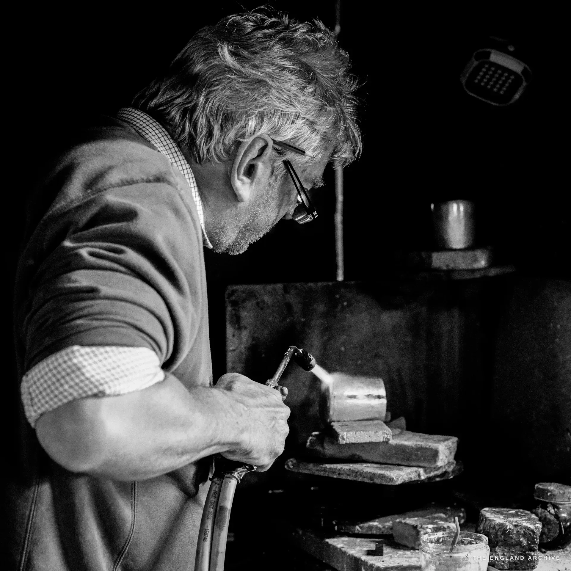 An elderly man in a checked-collar tank top works a silver blank on the stake with a small tool, an electrical box on the dark wall behind.