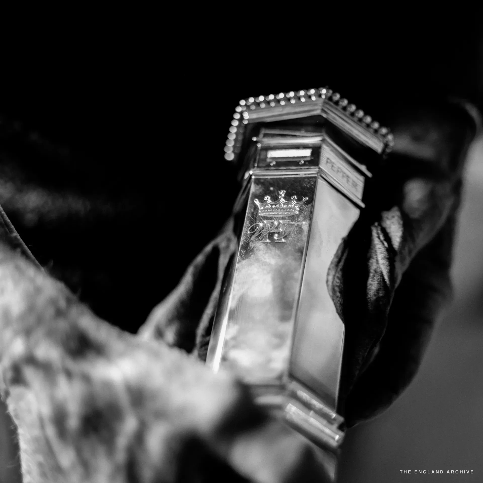 An extreme close-up of a hexagonal silver pepper caster engraved with a coronet above an entwined WH cipher, its lid pierced with beading.