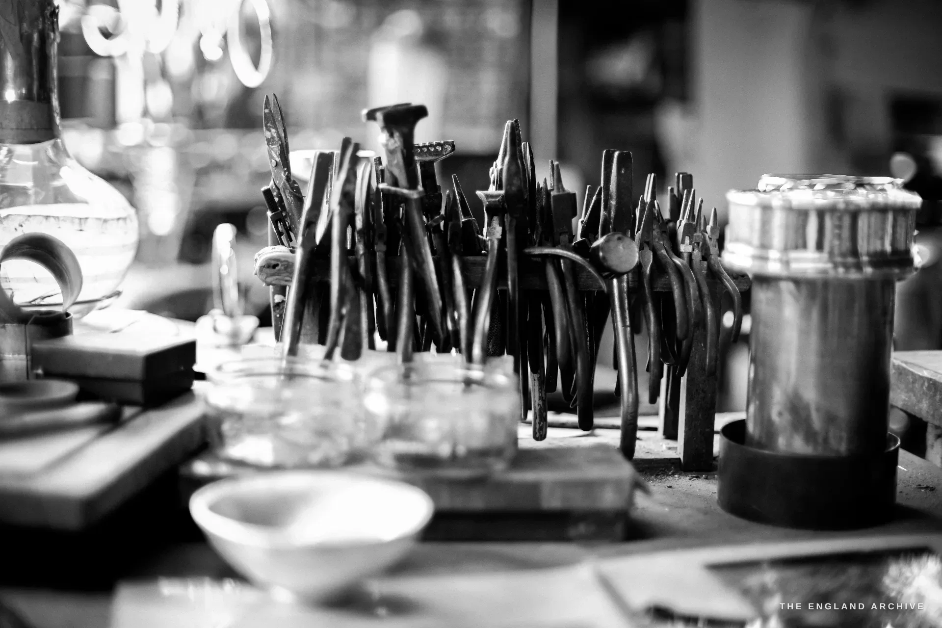 A dense bench-top thicket of dark-handled chasing and forming tools stood upright in a block, with glass jars and a small white crucible in front.