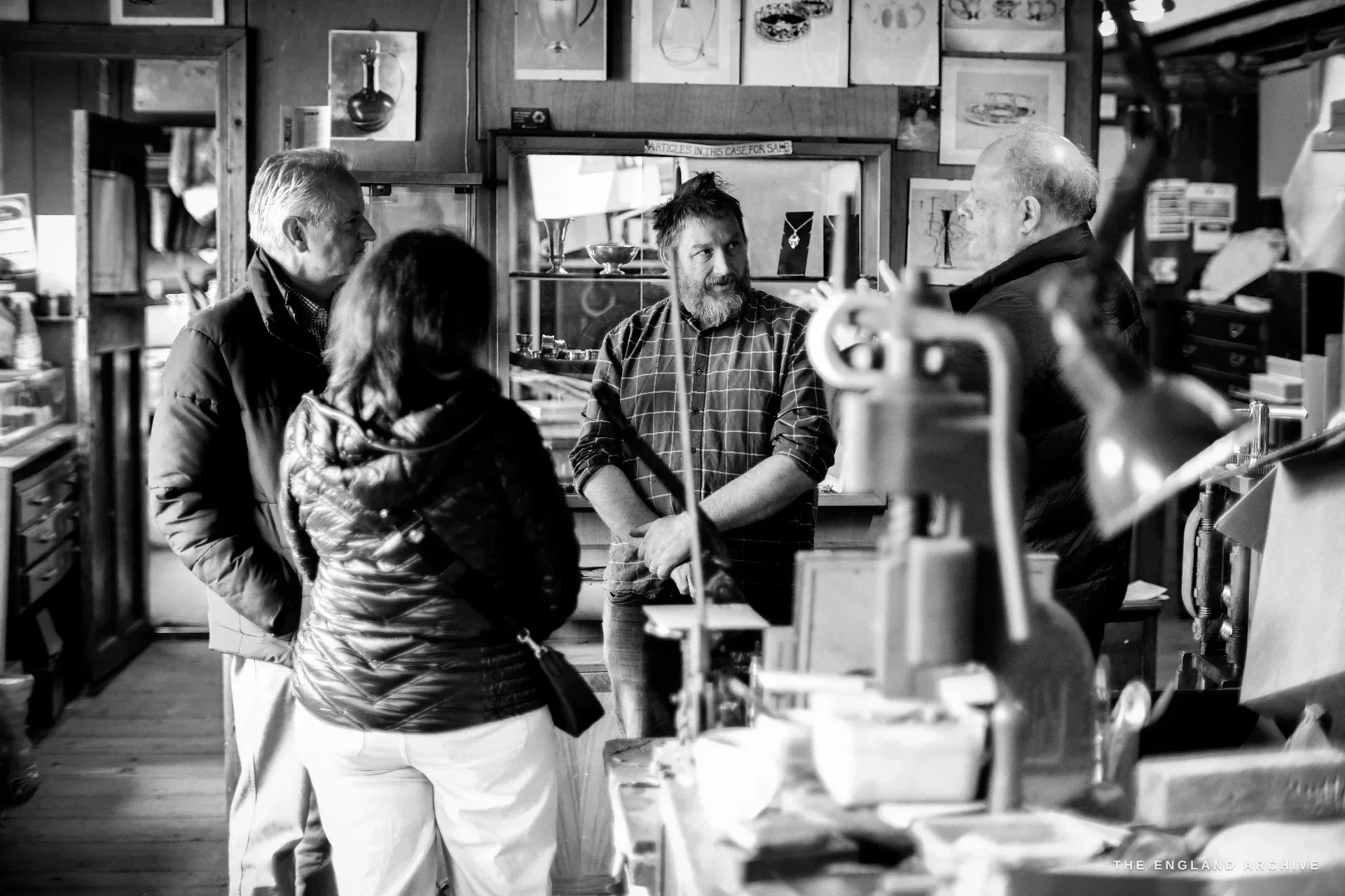 A bearded craftsman in a checked shirt talks with two visitors across a press in the lived-in workshop.