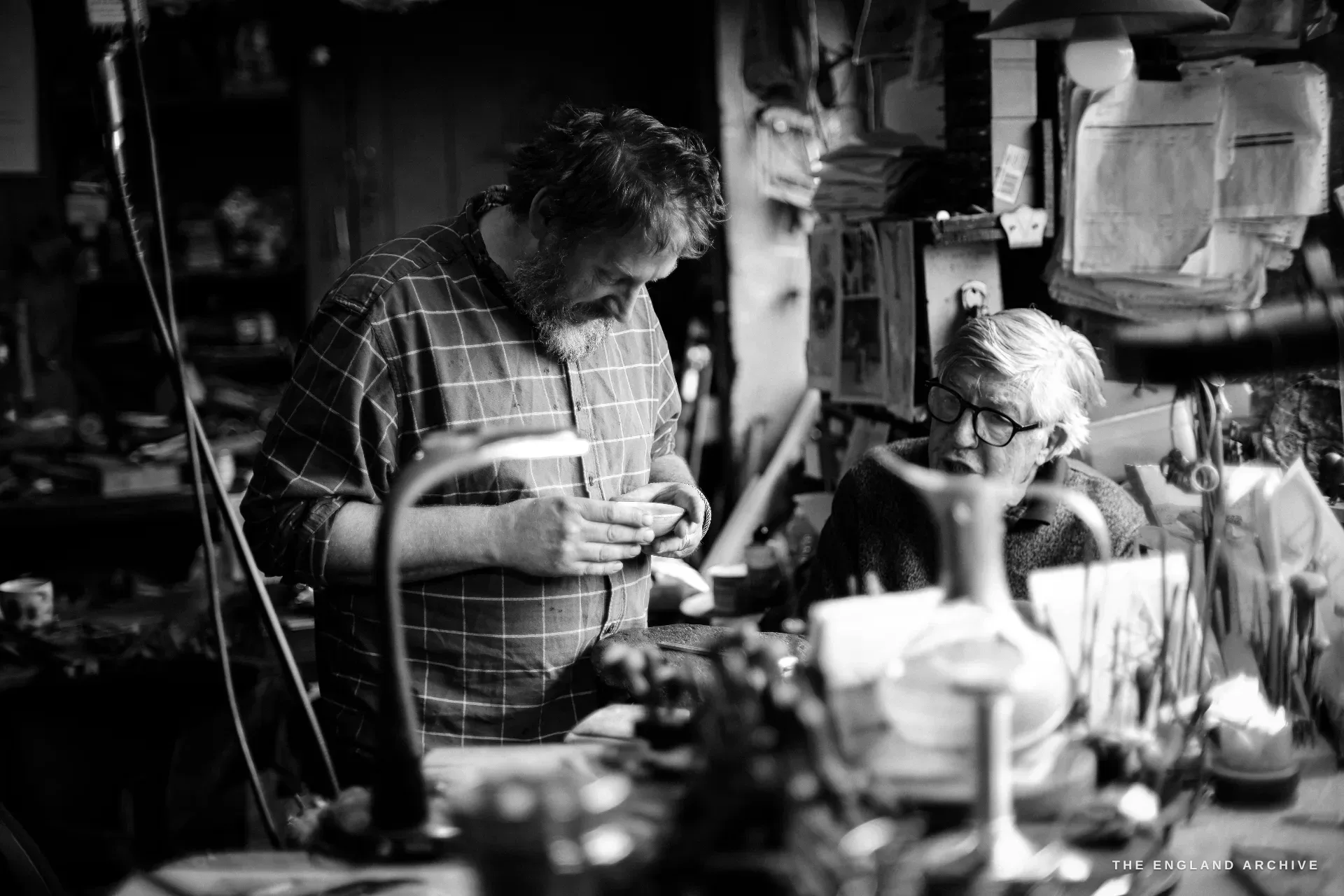 A bearded craftsman in a checked shirt works a small silver piece while David Hart looks on from behind in the cluttered workshop.