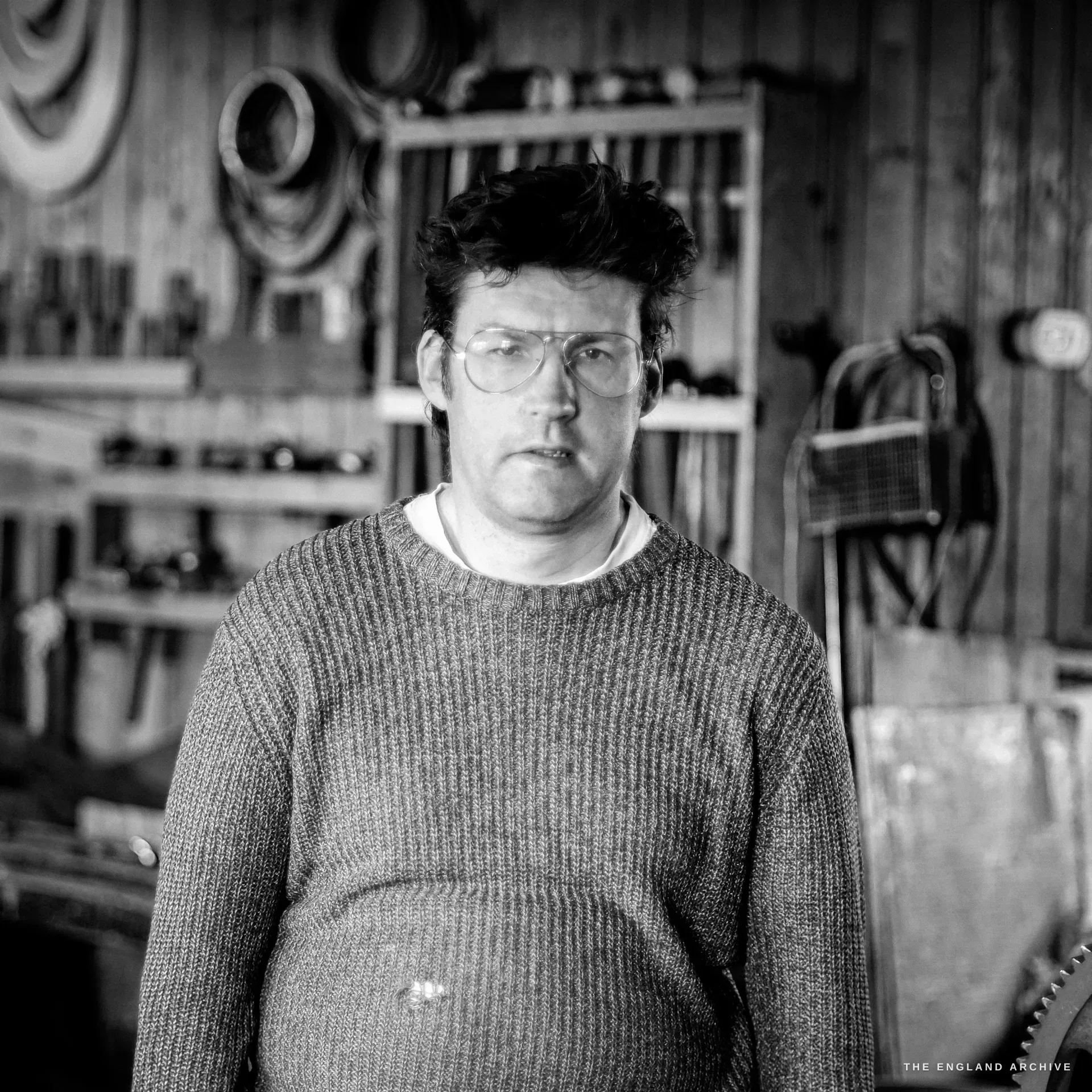 An environmental portrait of a heavy-set bespectacled craftsman in a grey jumper facing the camera, a wall of tools and a hand-saw behind him.
