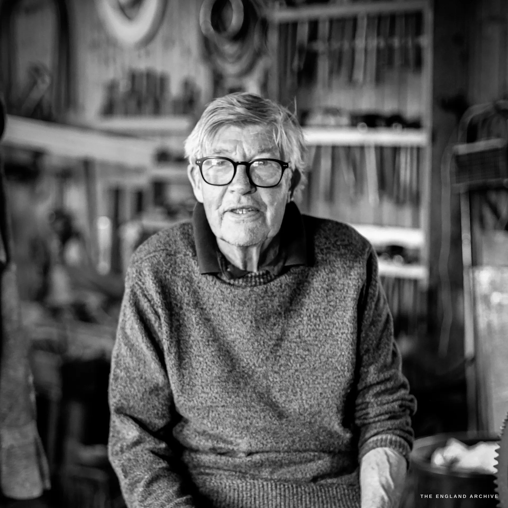 An environmental portrait of David Hart, white-haired in black-framed glasses and a grey jumper, seated and addressing the camera amid his tool racks.