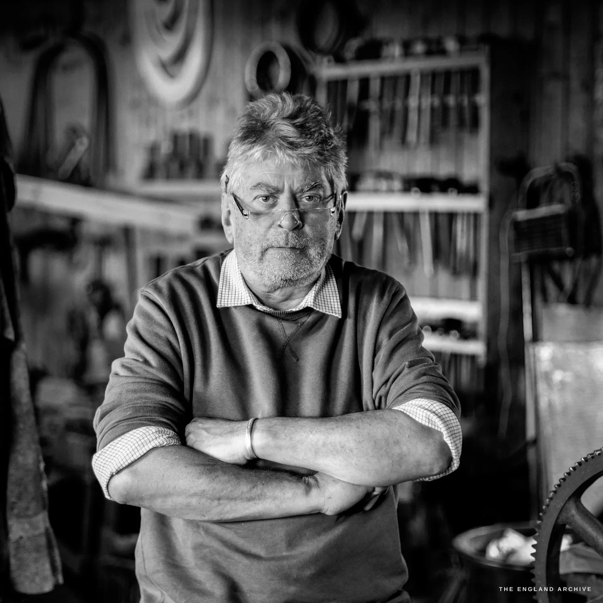 An environmental portrait of a grey-haired bespectacled craftsman, arms folded, in a tank top over a checked shirt, before the workshop tool wall.