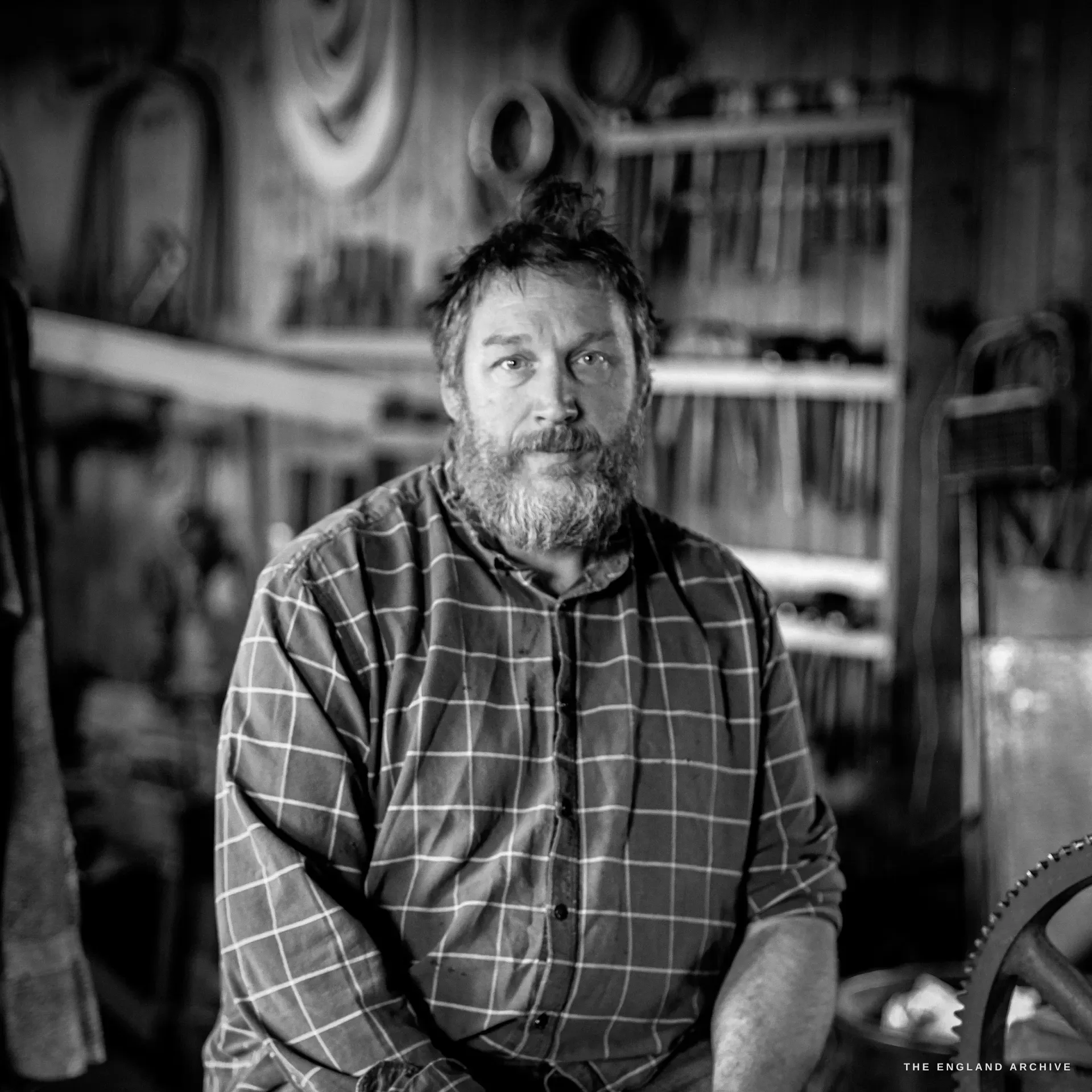 An environmental portrait of a bearded man with his hair in a top-knot, in a checked shirt, seated with arms resting, the workshop's hanging tools behind him.