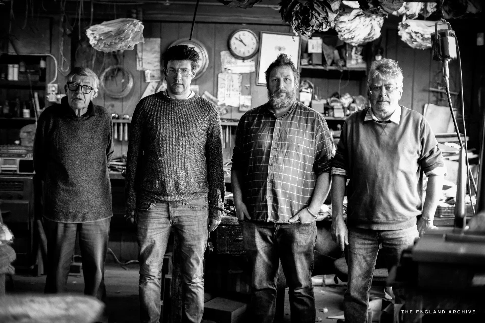 A group portrait of four Hart's silversmiths lined up across the workshop, the elderly David Hart at the left, a wall clock and hanging dockets above them.