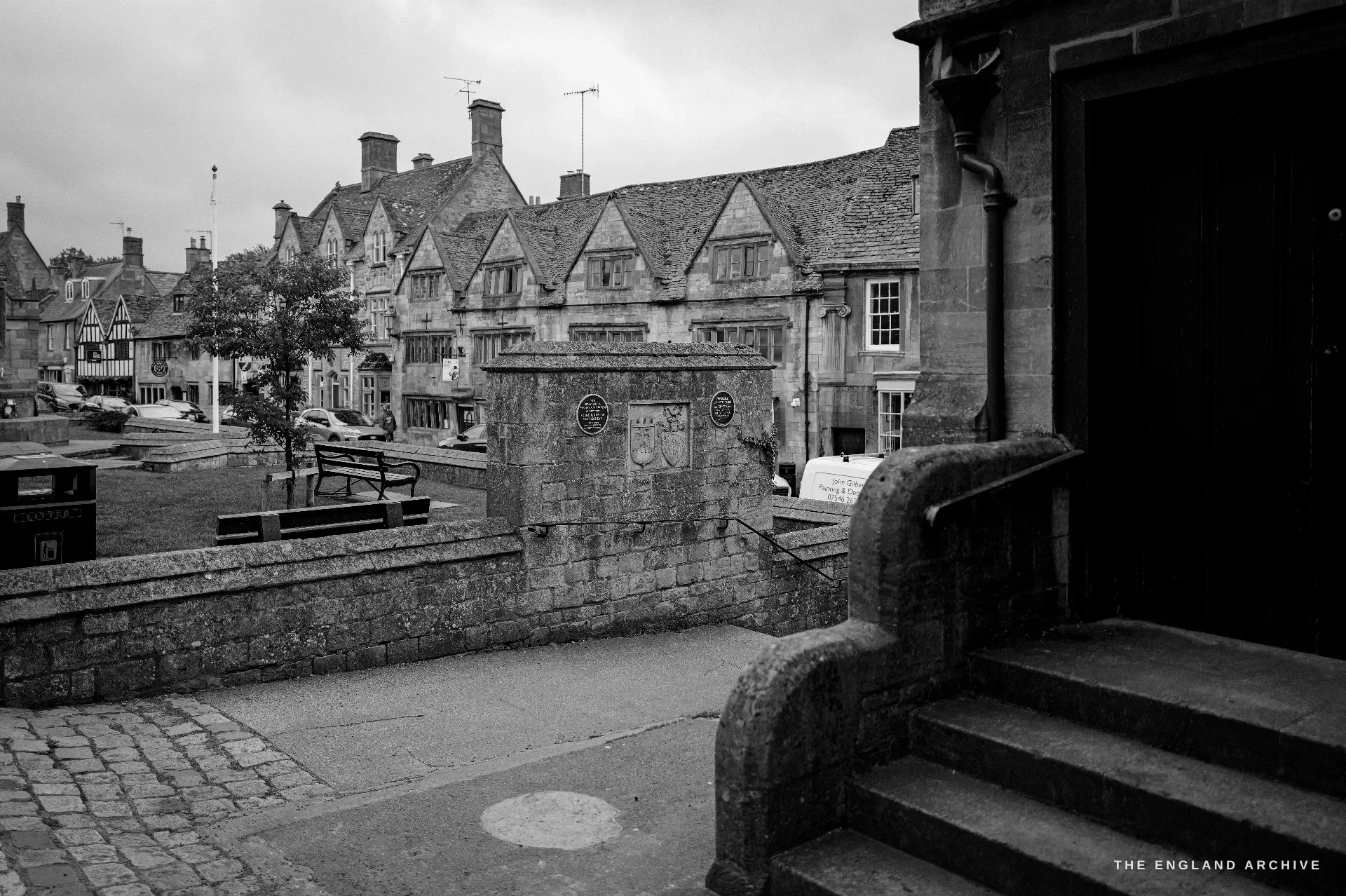 Chipping Campden High Street seen from beside stone steps, past the railed Cotswold-stone war memorial fountain to a long terrace of gabled limestone houses under a flat grey sky.
