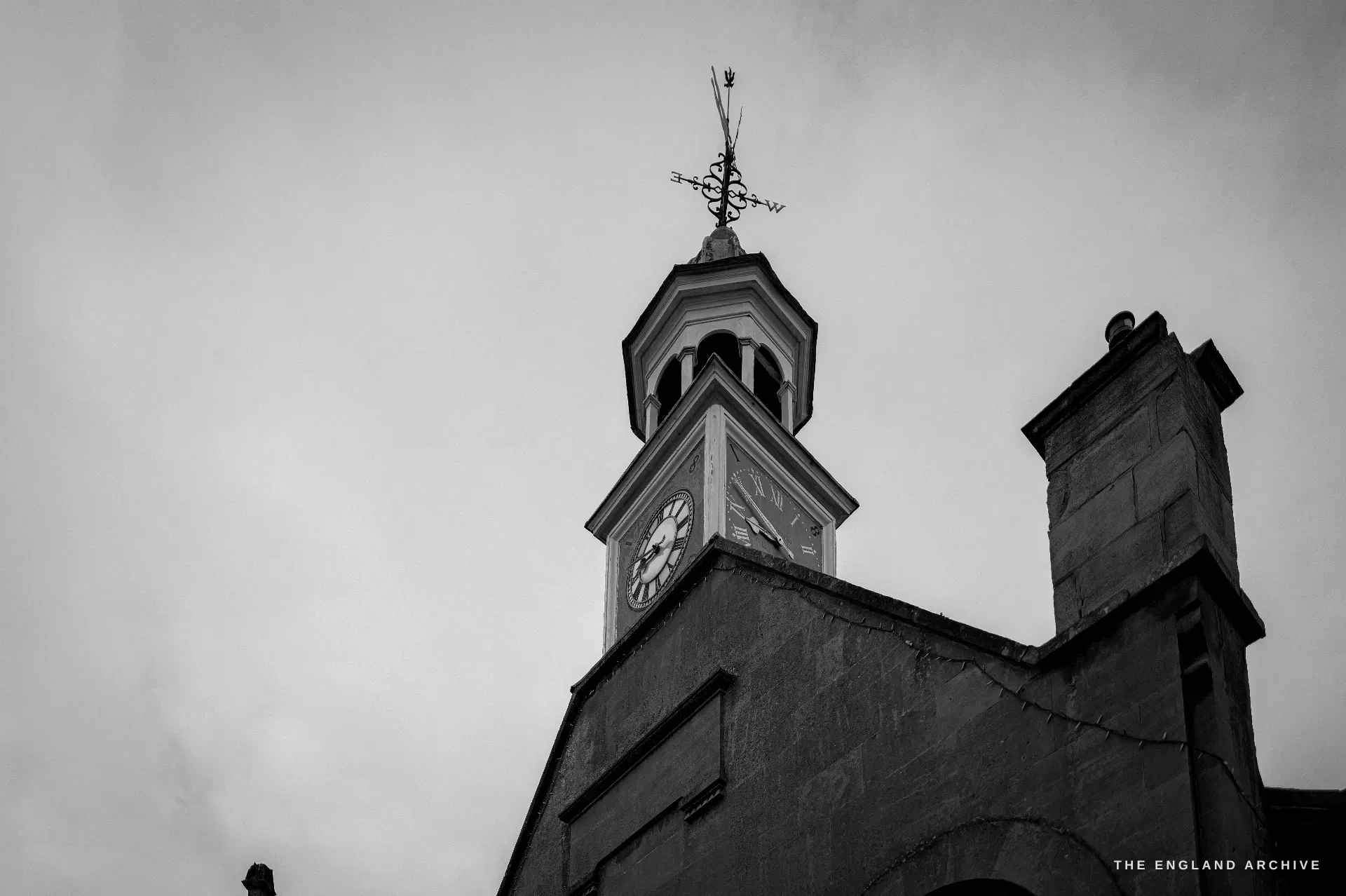 An upward study of the Market Hall bell-cupola and clock against cloud, a wrought-iron weathervane spiking off the lead-roofed lantern above a stone gable.