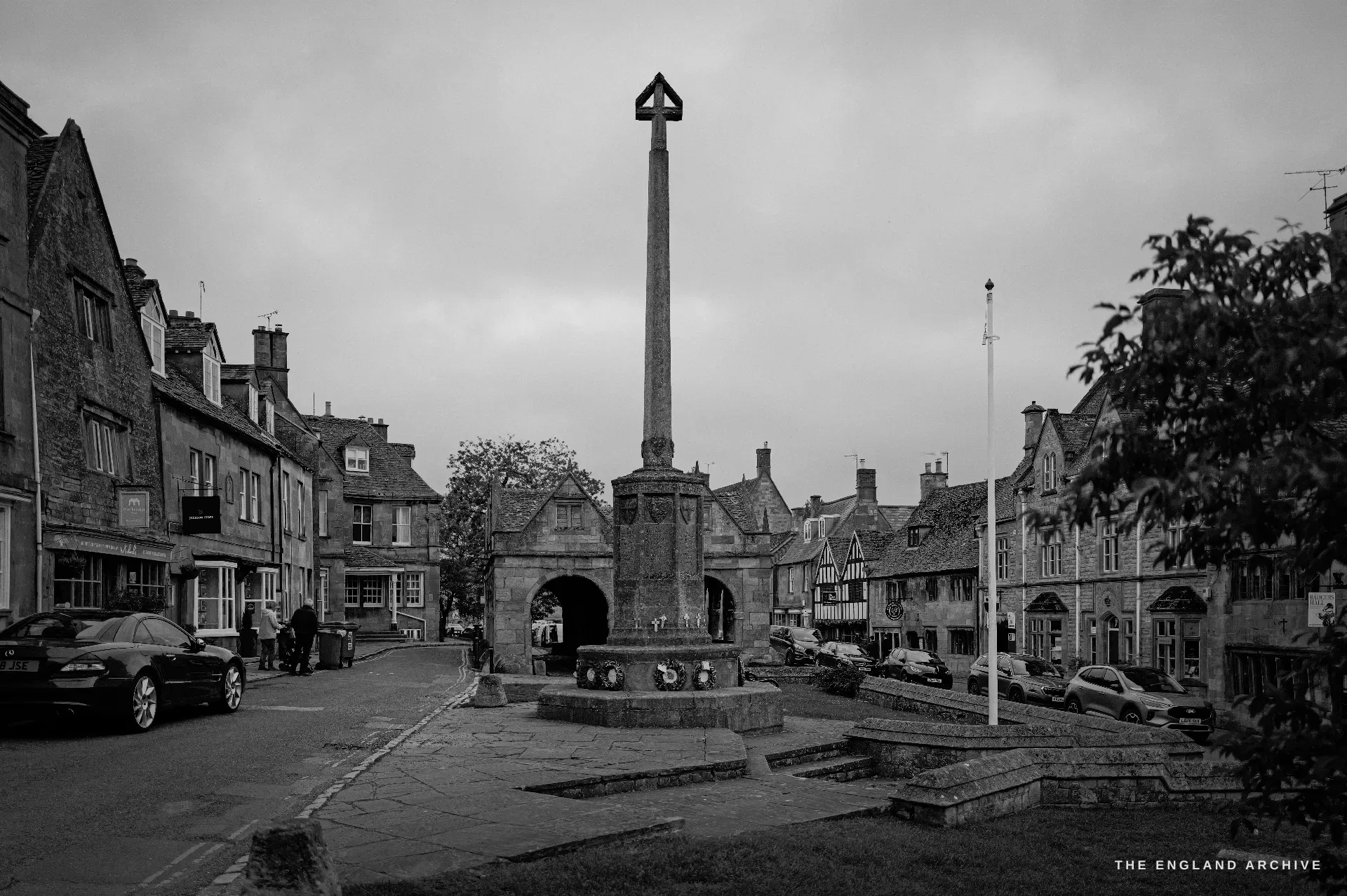 The medieval High Cross stands on its stepped octagonal base in the middle of the street, framed by parked cars and the gabled stone frontages of the High Street.