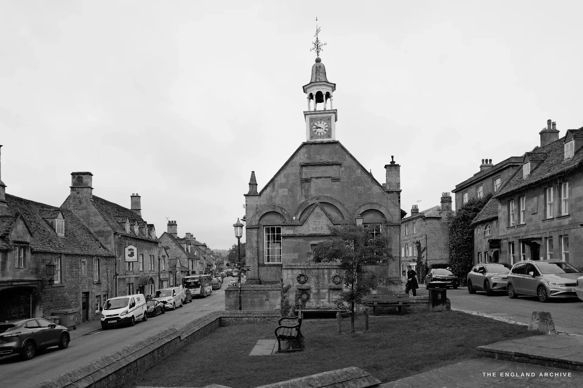 A wider street view of the gabled Market Hall with its bell-cupola and clock, flanked by Cotswold cottages and parked cars down the High Street.
