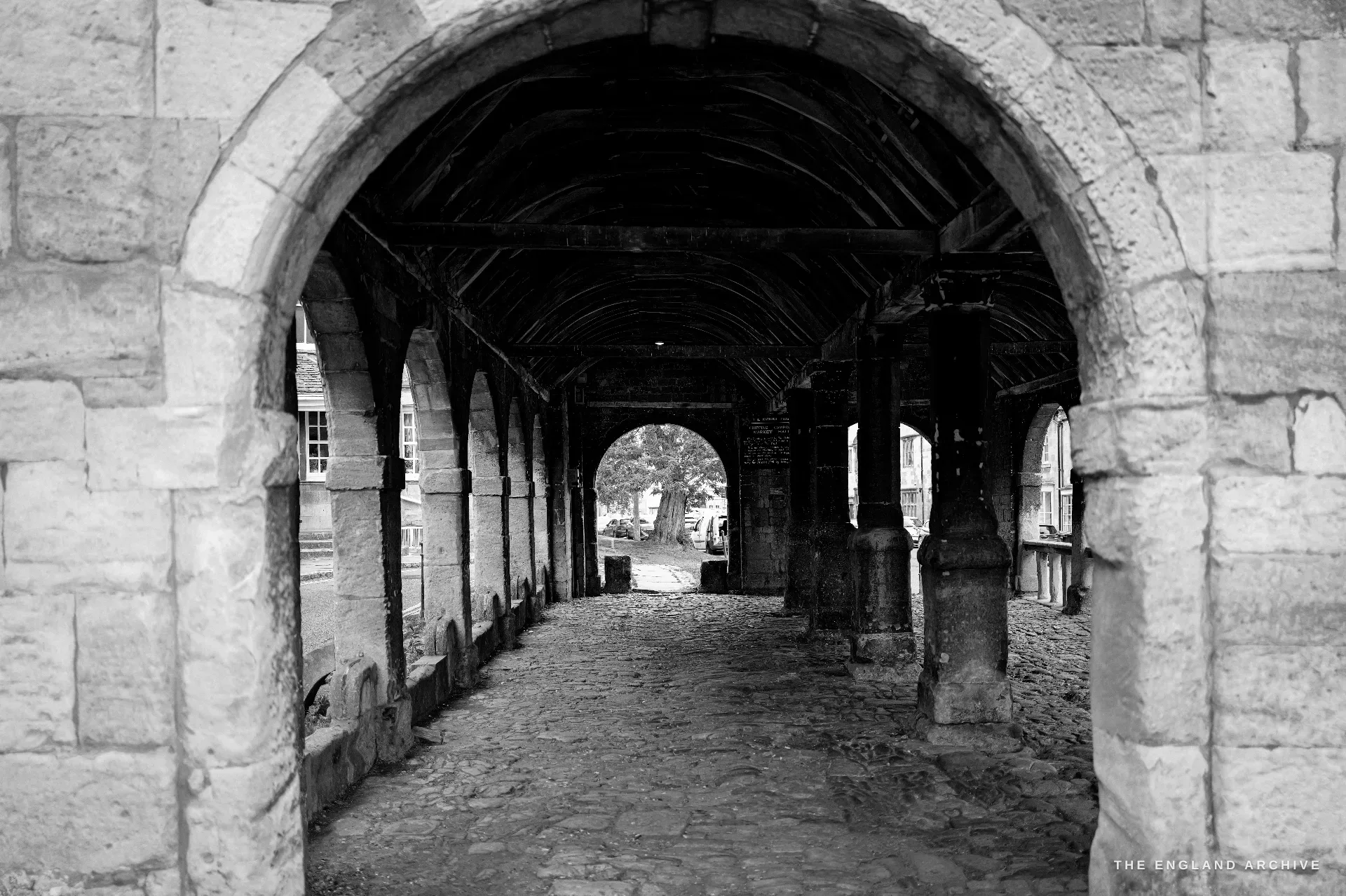 Looking down the worn flagstone aisle of the Market Hall arcade under its timber roof, the stone arches framing the bright street beyond.