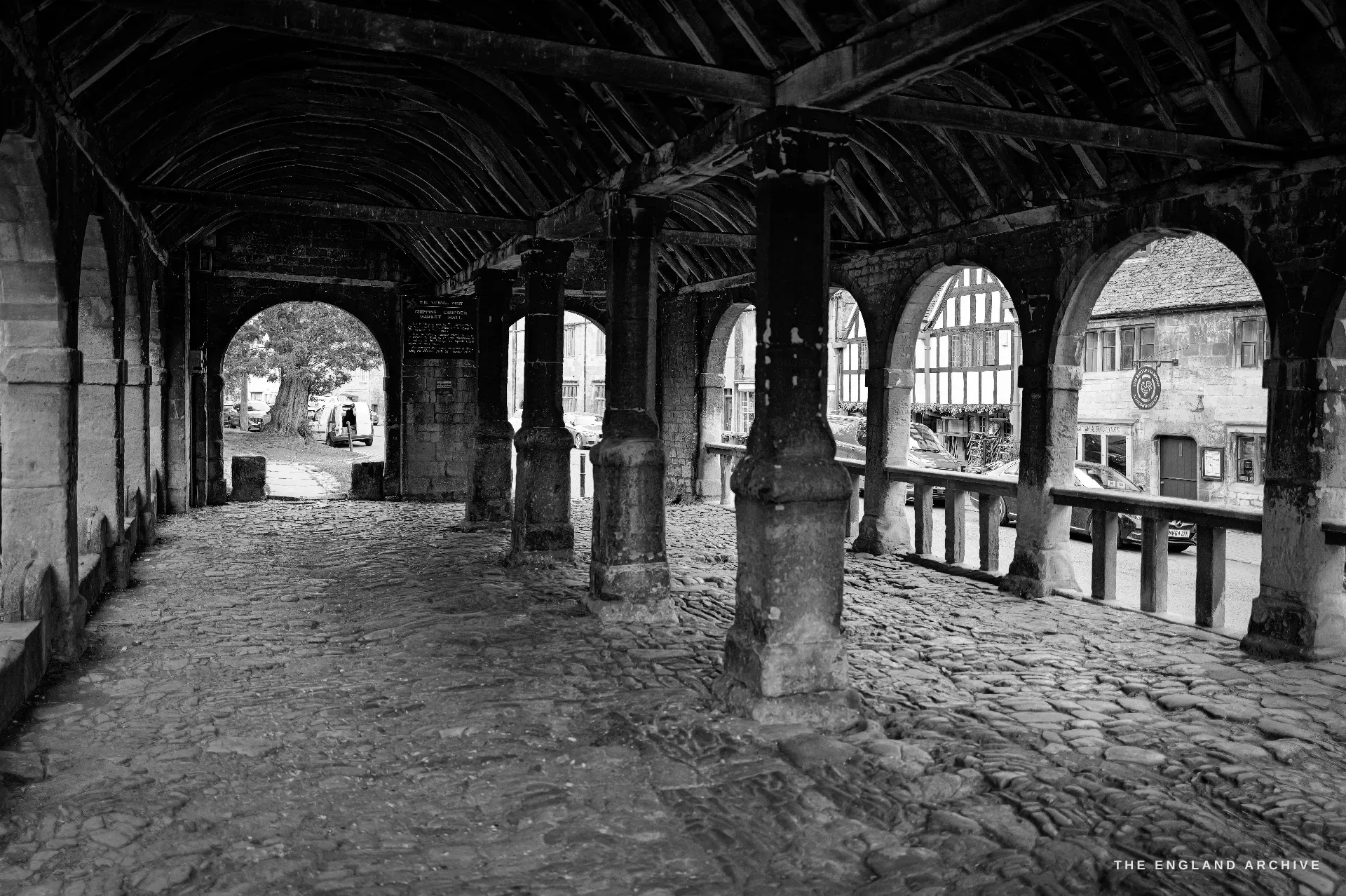 The Market Hall arcade from the corner, stone piers and round-headed arches over a cobbled floor, a half-timbered building visible through the openings.