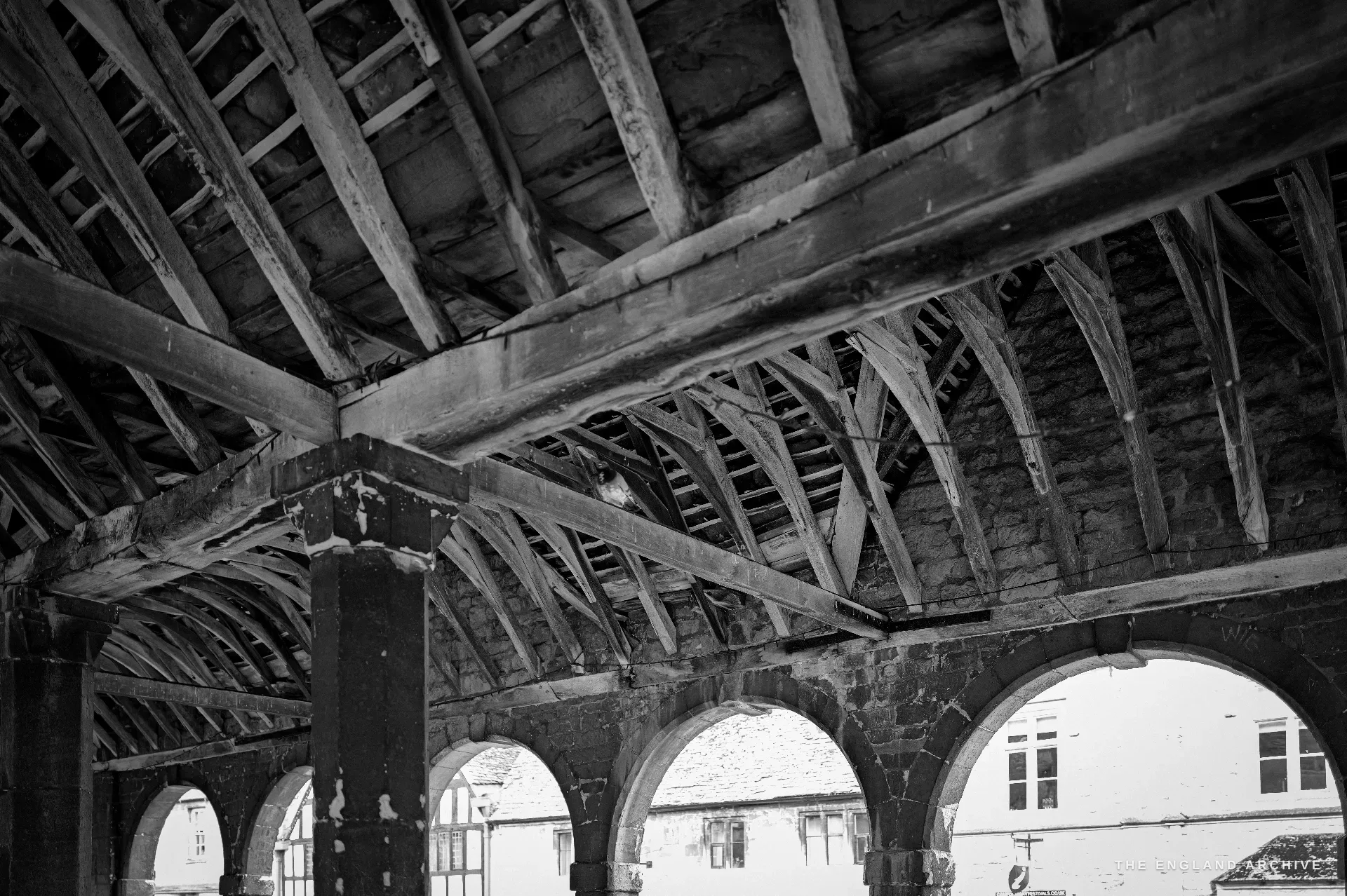 The Market Hall's heavy timber roof beams and curved braces seen from inside, a run of stone arches and the bright street along the lower edge.