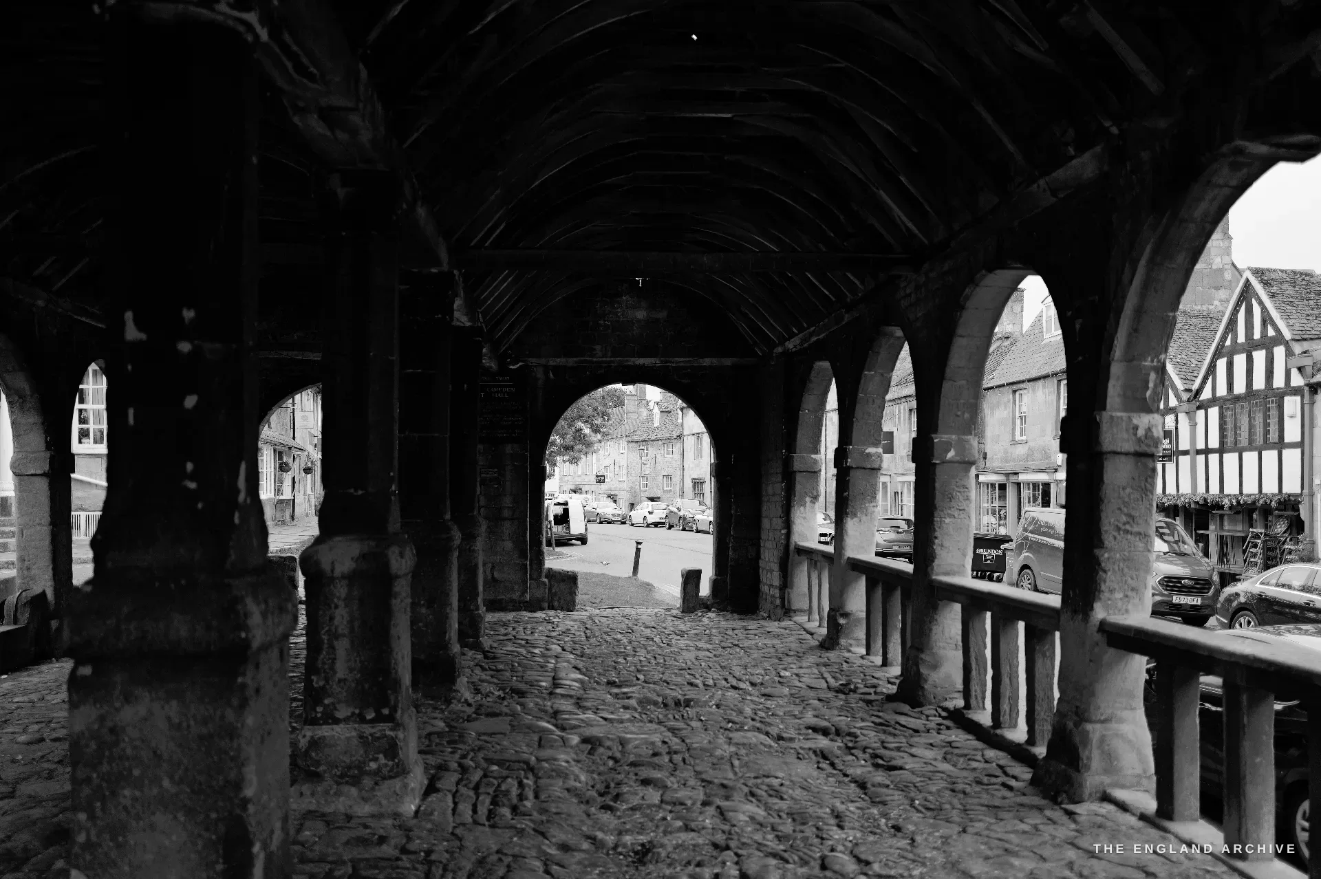 A view out from within the Market Hall arcade, a central pointed arch framing parked cars and a half-timbered house across the street.